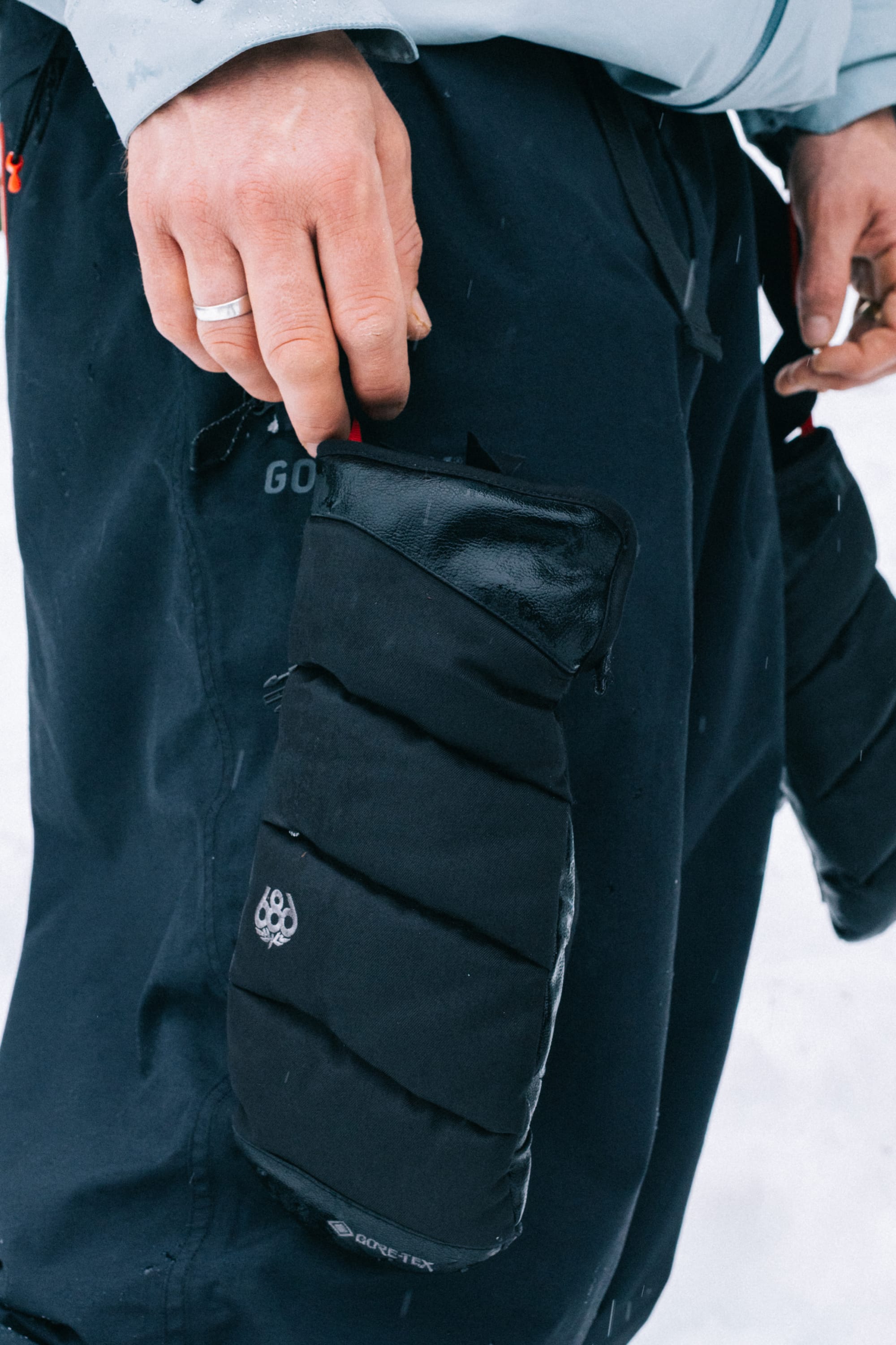 A person wearing dark snow pants with a 686 Men’s GORE-TEX® Buffer Insulated Mitt by 686 attached to their leg stands against a snowy background, a ring visible on their left hand.