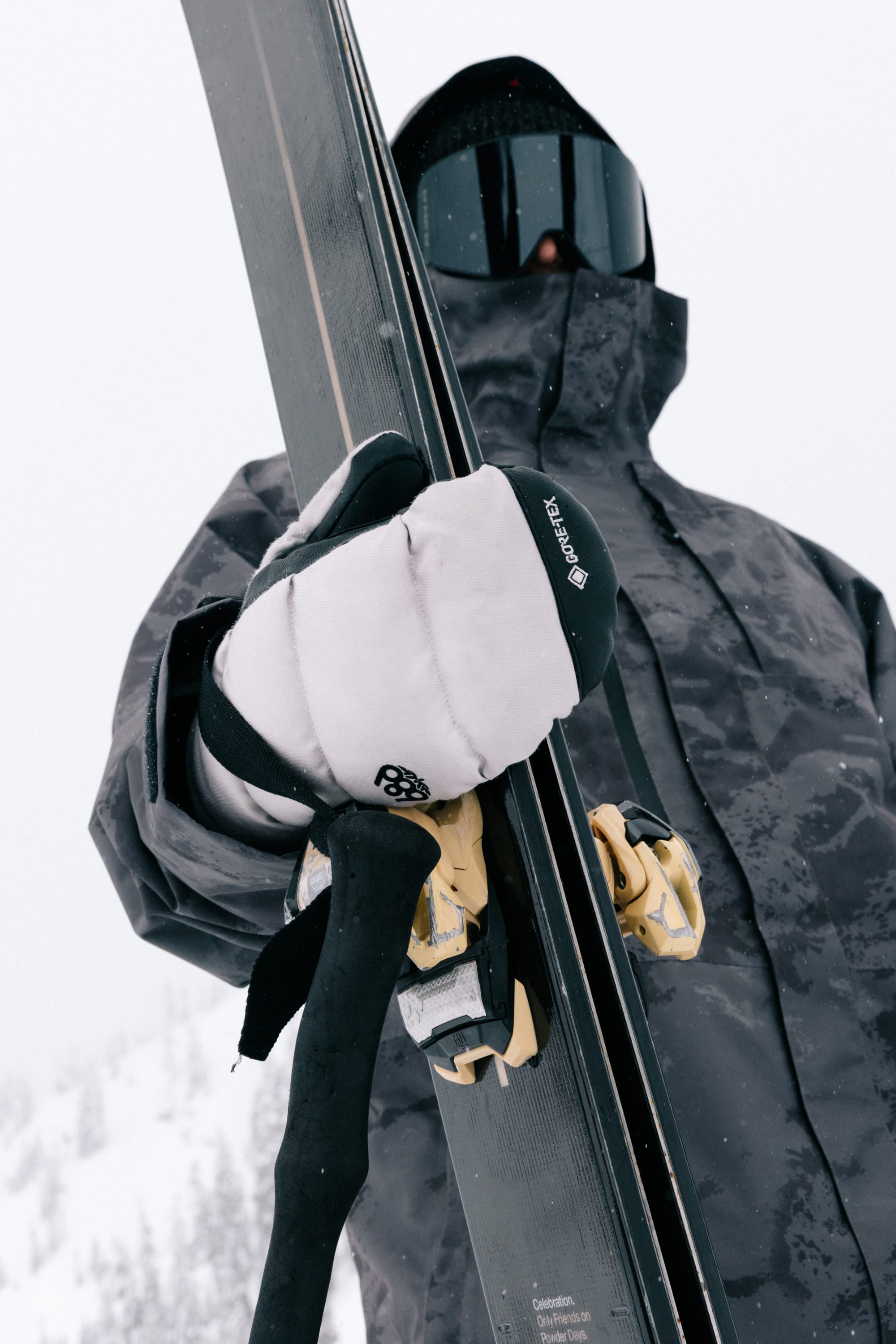 A skier in a gray jacket, 686 Men’s GORE-TEX® Buffer Insulated Mitts by 686, and black goggles holds skis and poles on a snowy mountain. The skier’s face is mostly covered, with snow-covered trees visible in the background.