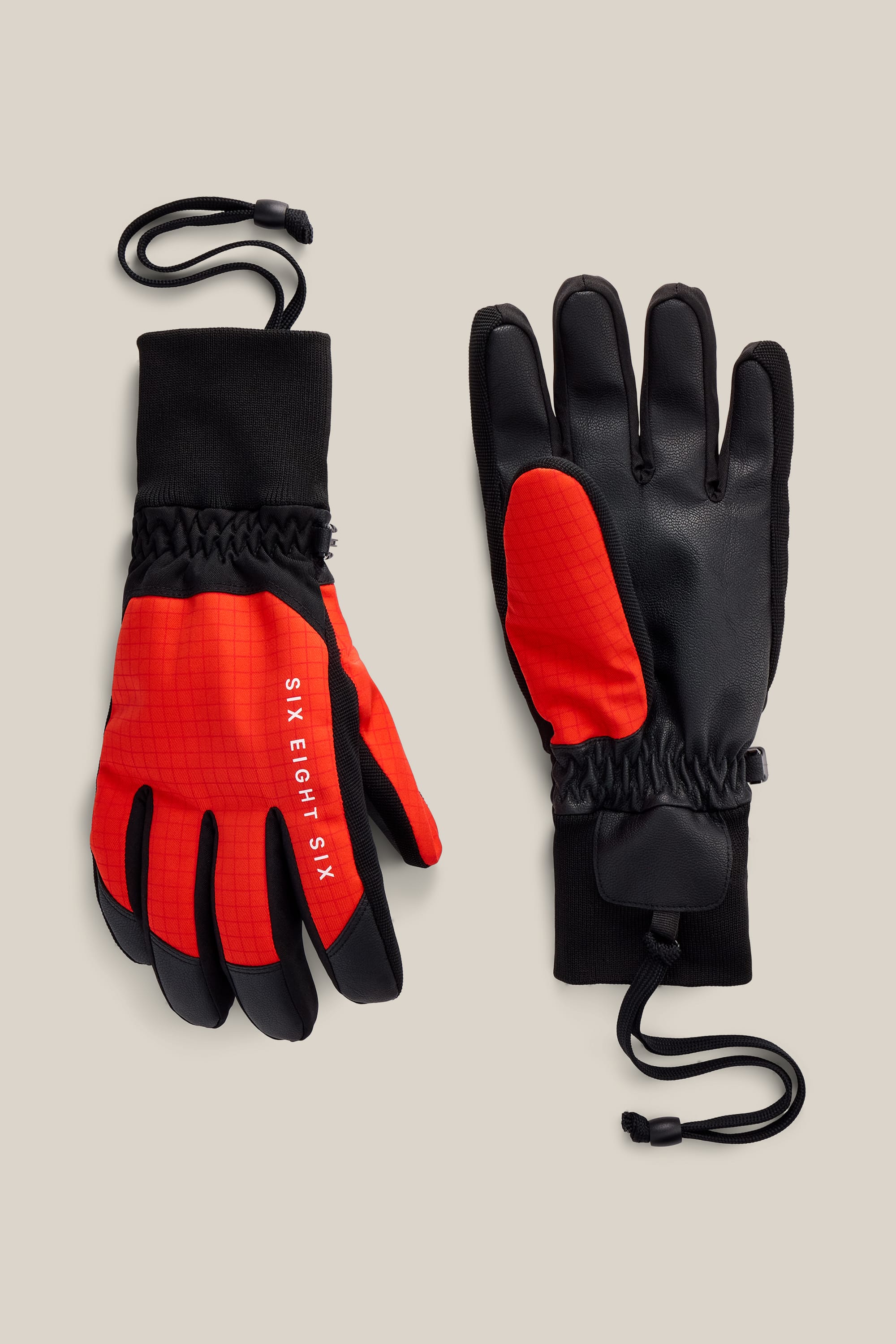 A pair of 686 Men’s Crush Insulated Gloves in black and red, featuring wrist cinches and adjustable cords from 686, displayed palm-up and palm-down on a beige background.
