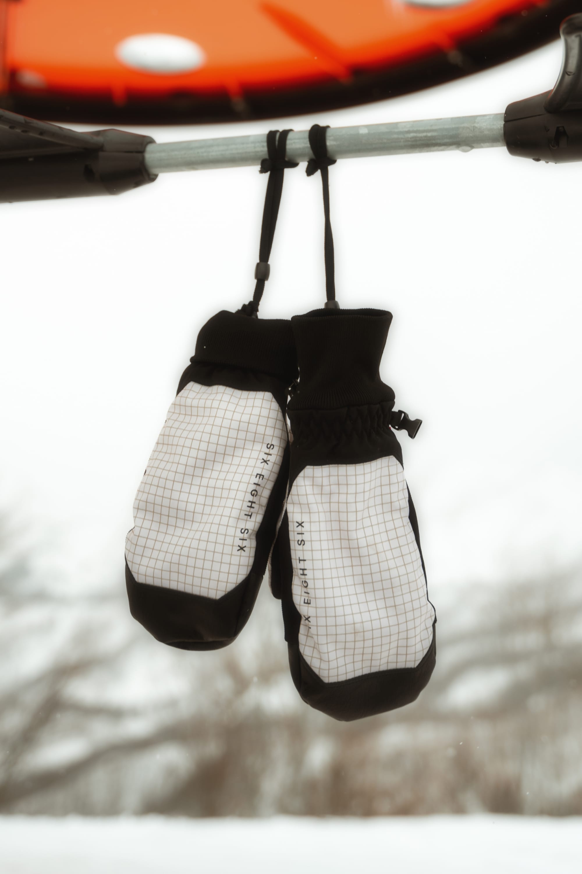 A pair of 686 Men’s Crush Insulated Mitts in a black and white grid pattern hang from a metal bar, with a snowy outdoor background softly blurred behind them.