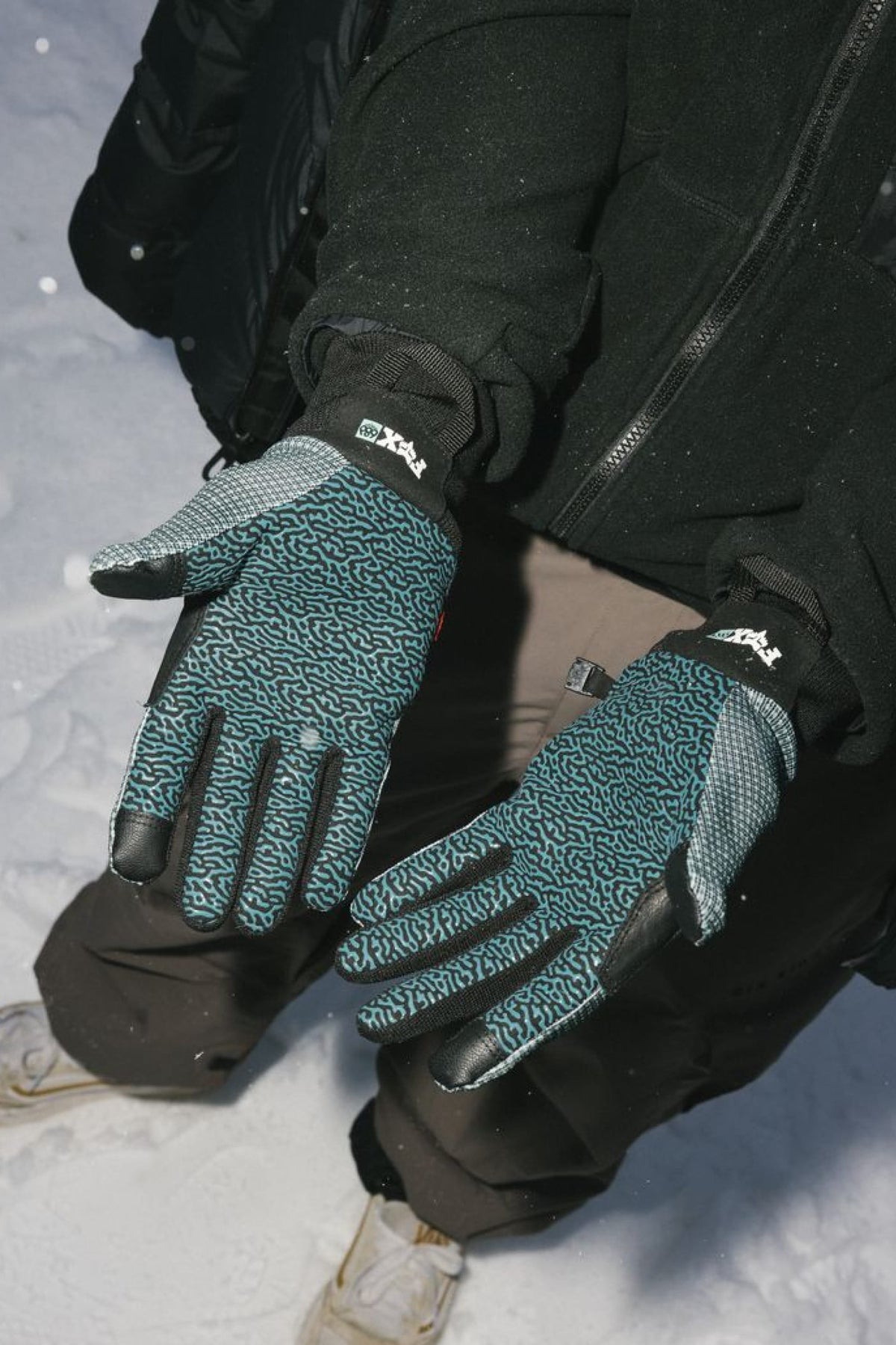 Wearing the 686 Men’s Hold Up Pipe Glove and a black jacket, this person stands on snow. Only their gloved hands, jacket, brown pants, and white shoes are visible.