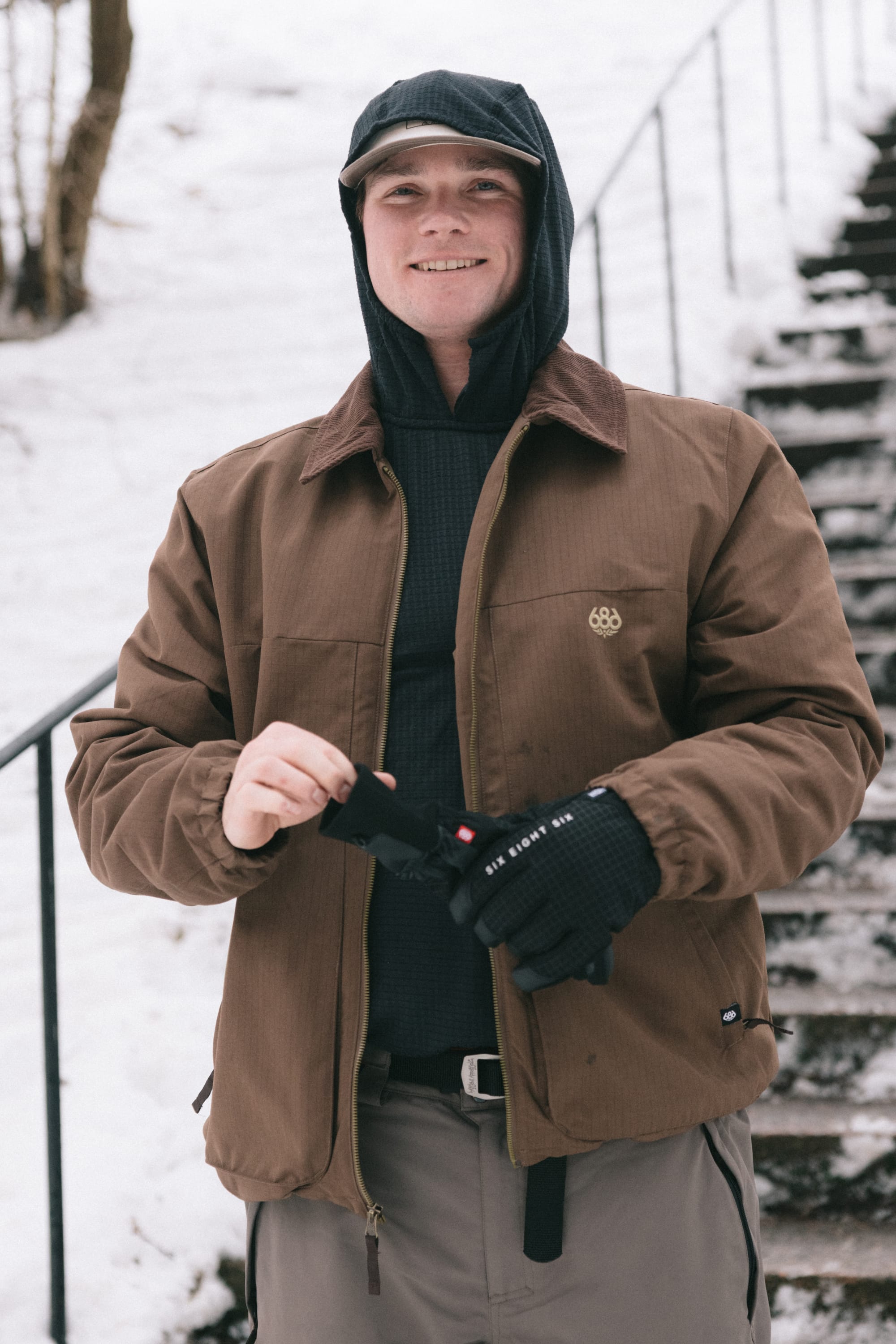 A person wearing the 686 Men’s 686 Camber Insulated Jacket in brown by 686, paired with black gloves and a hood, stands outside on a snowy day near steps, smiling at the camera.