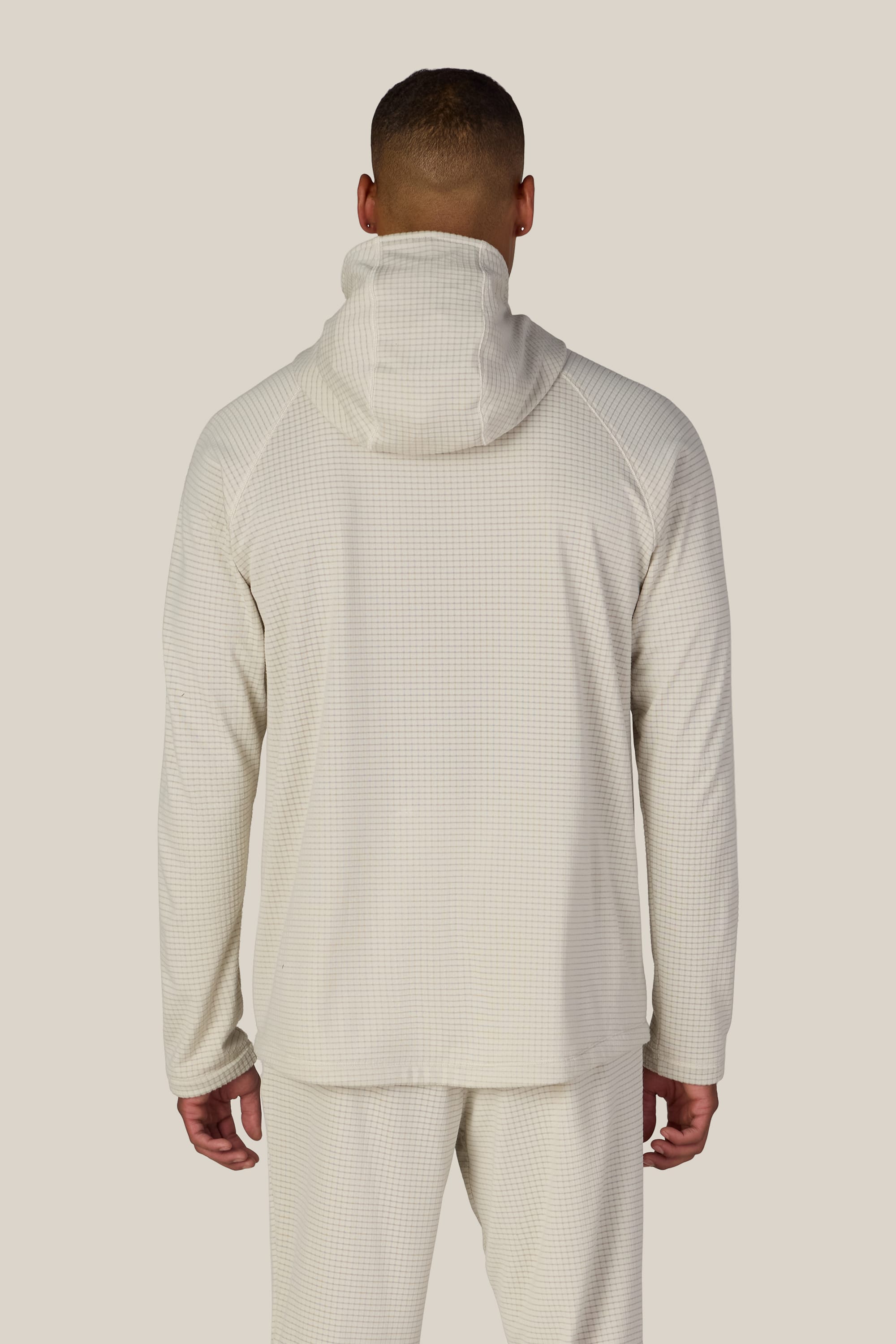 A person stands facing away, wearing the 686 Men’s Mission Grid™ Raglan Hoody in a light color with matching pants. The outfit showcases a subtle checkered pattern, enhancing its contemporary style against a plain, neutral background.