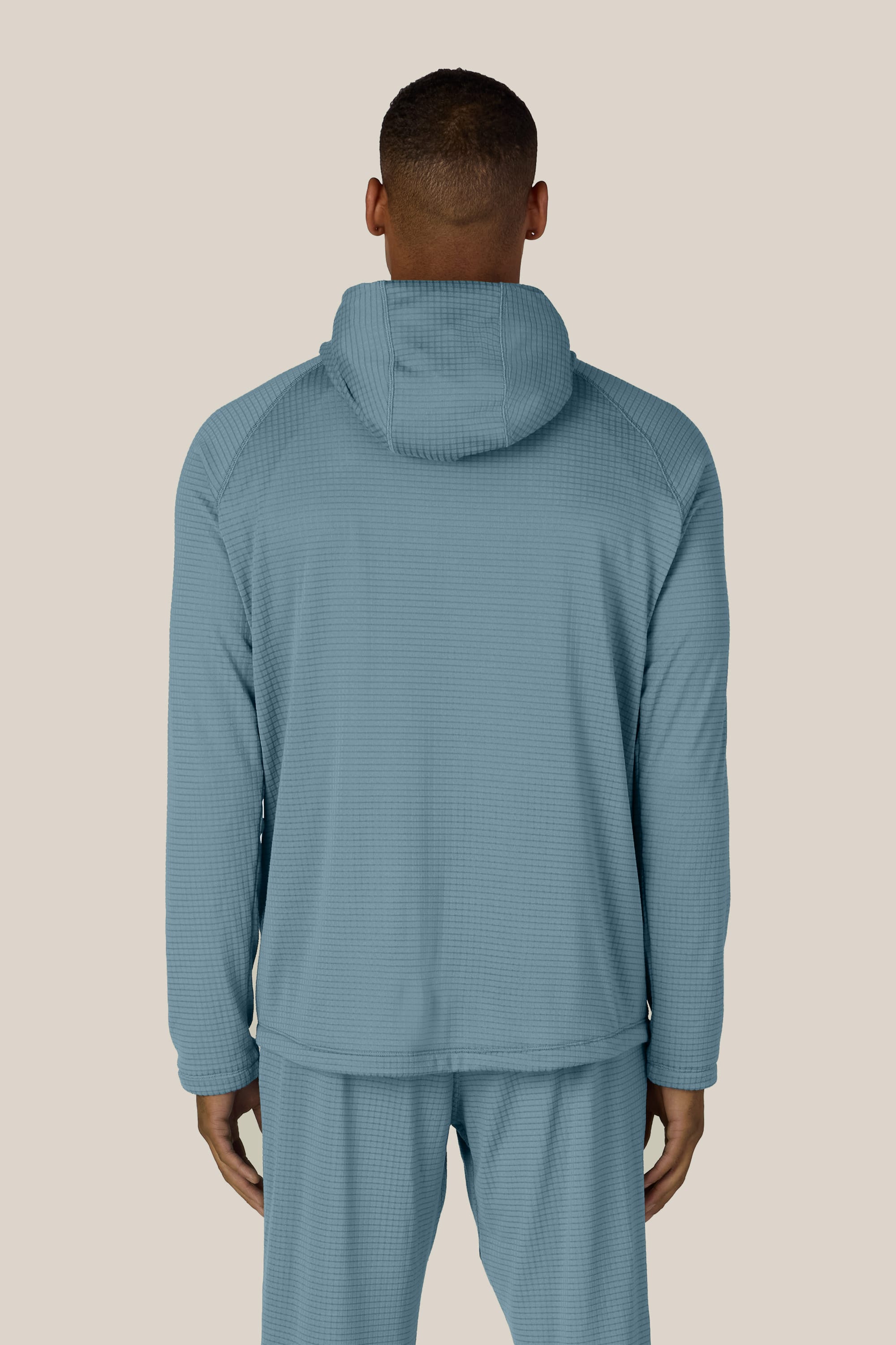 A person stands facing away, wearing the 686 Men’s Mission Grid™ Raglan Hoody in light blue and matching pants against a plain beige background.
