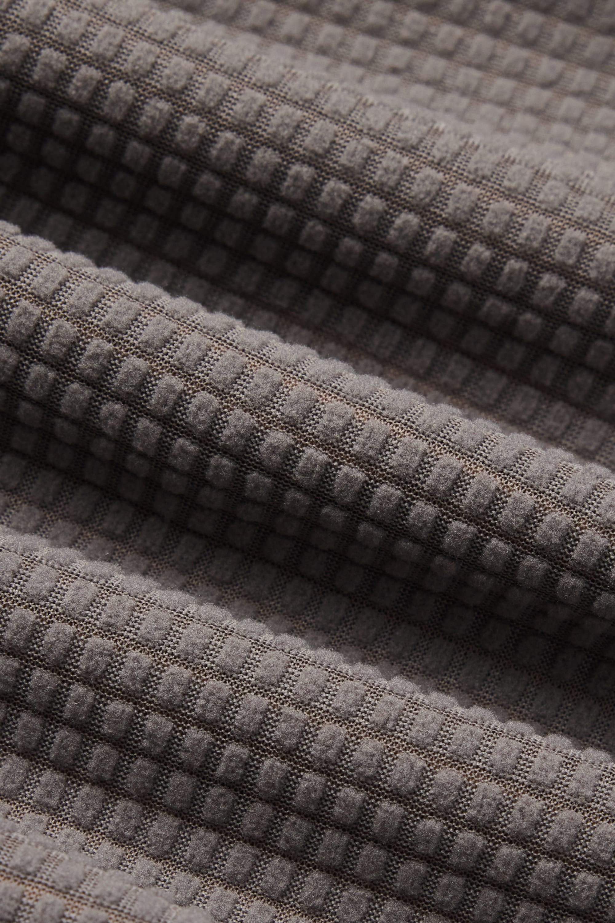 Close-up of the 686 Men’s Mission Grid™ Pant fabric in gray, featuring a waffle-textured grid pattern with visible soft folds that highlight its three-dimensional surface and fine weave details—ideal for stylish men’s knitwear from 686.