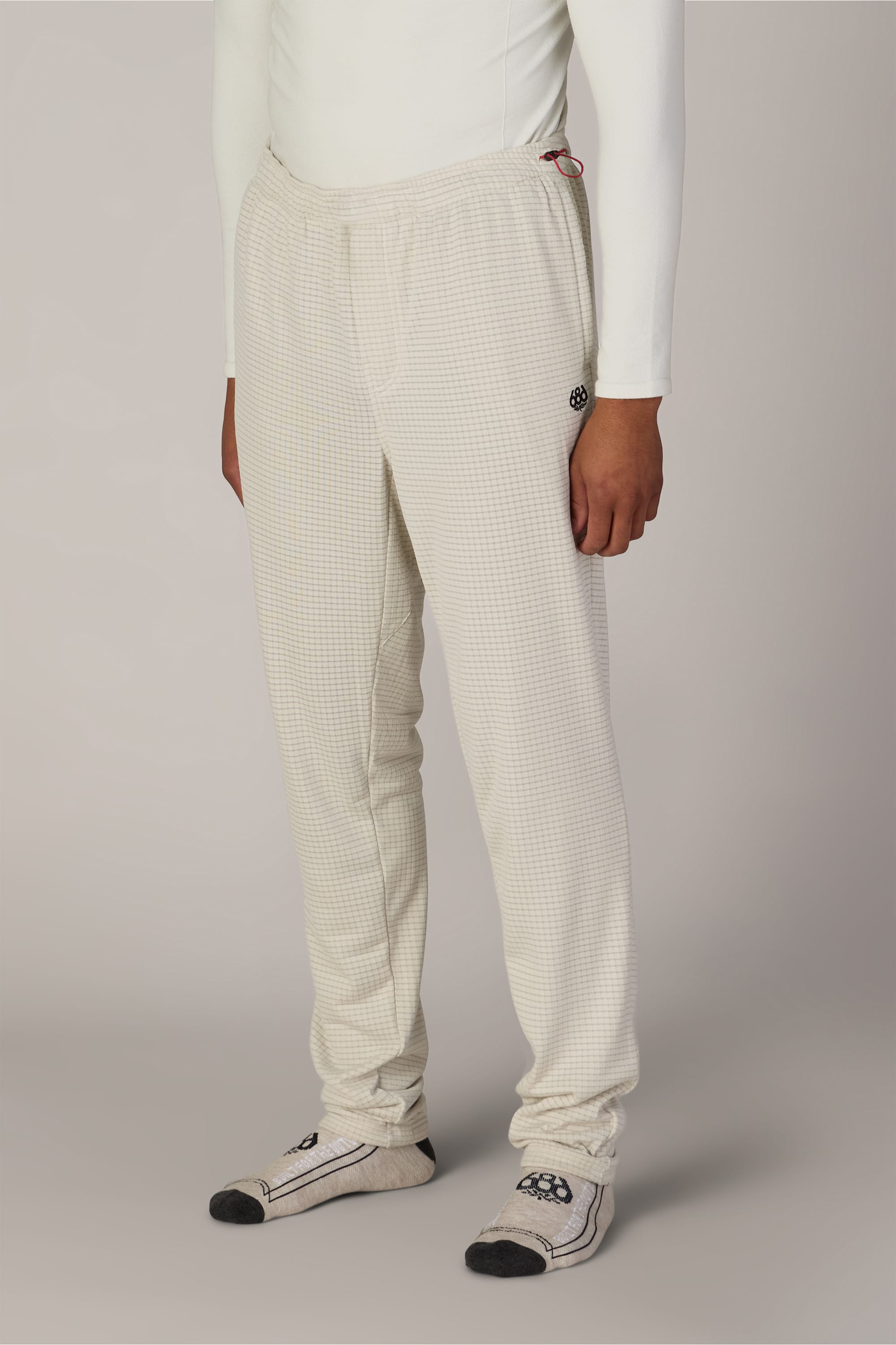 A person wearing a white long-sleeve top, the 686 Men’s Mission Grid™ Pant by 686—featuring an off-white knit design and elastic waistband—and gray socks with black toe and heel accents, standing against a plain light background.