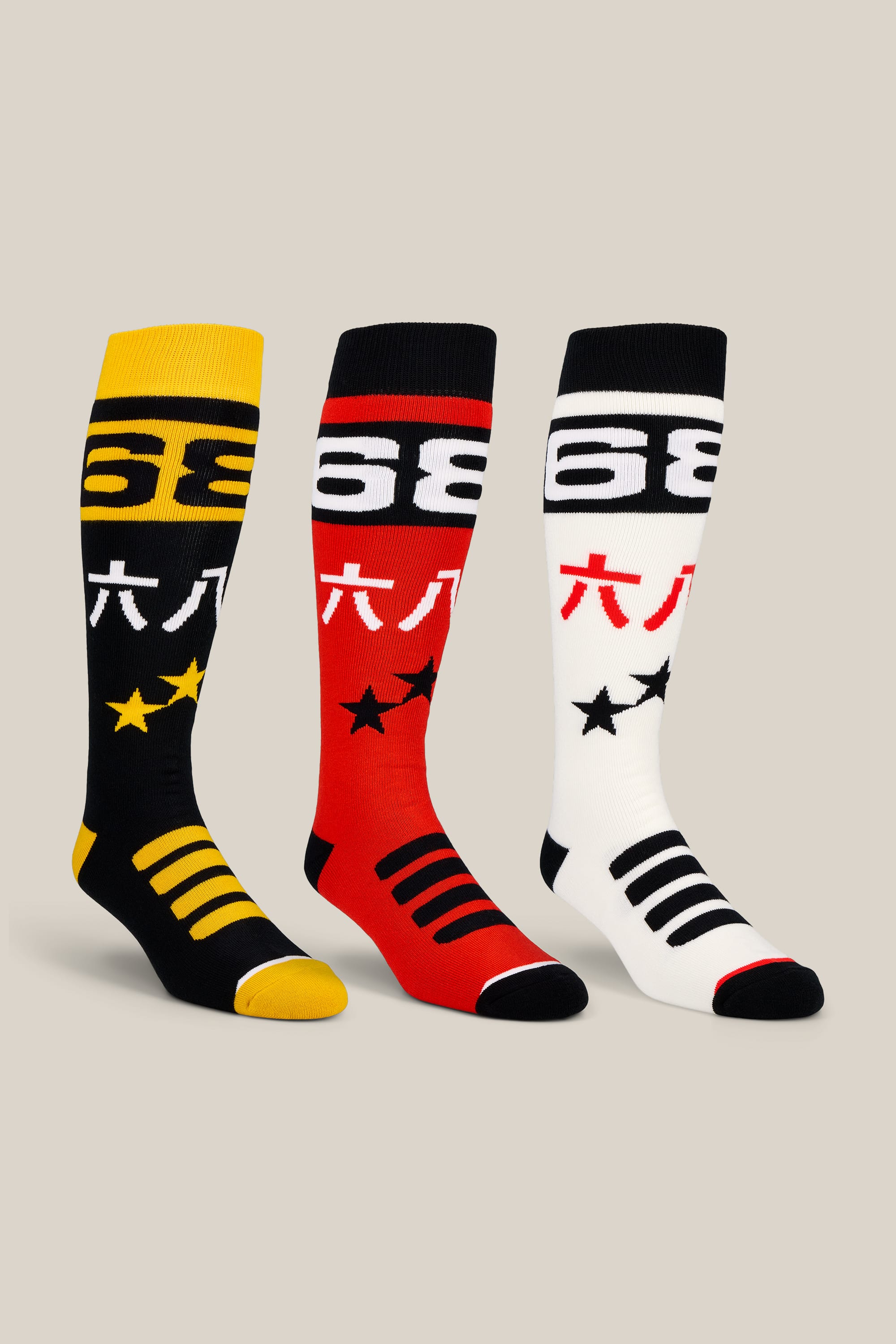 The 686 Men’s Kanji Socks - 3-Pack by 686 showcases three knee-high performance socks in yellow, red, and white, each featuring black accents, prominent stars, bold numbers, and Asian characters near the top. Displayed upright on a light beige background, these men’s socks are crafted for durability and comfort—ideal for adding a standout touch to any wardrobe.