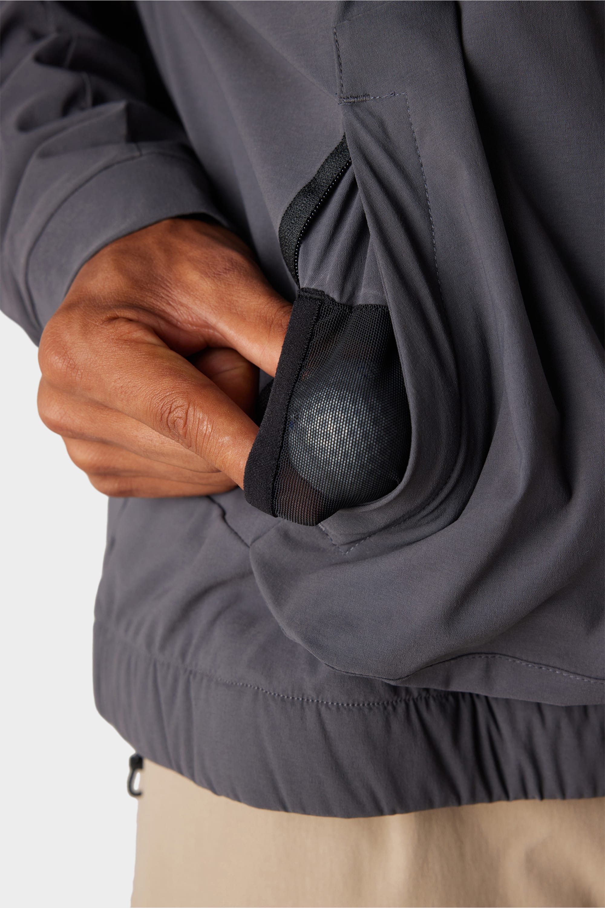 A hand holds a metallic ball in a mesh-lined pocket of the 686 x Malbon Mens PrimaLoft drirelease® Merino-Lined Pullover Jacket, grey with an elastic hem, paired with beige pants.