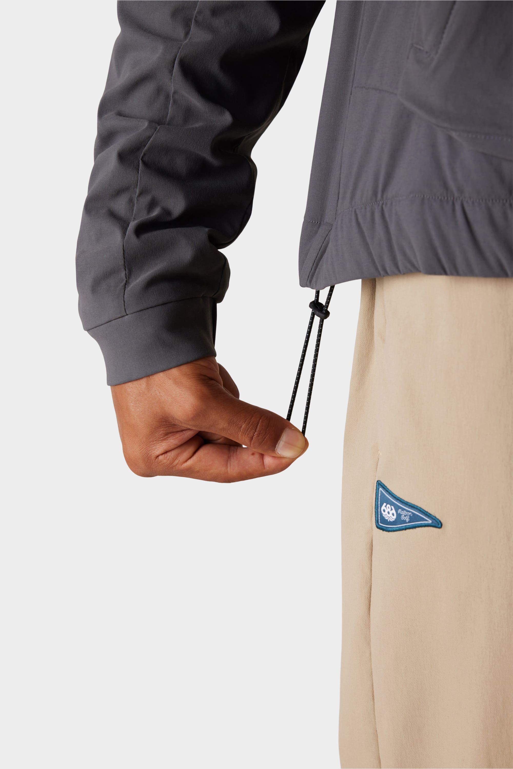 A person adjusts the drawstring hem of the 686 x Malbon Mens PrimaLoft drirelease® Merino-Lined Pullover Jacket in grey, paired with beige pants featuring a blue and white patch, set against a plain white background.