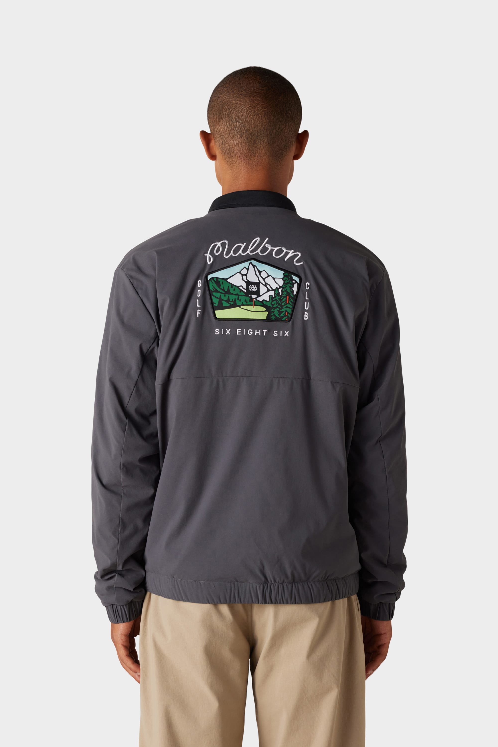 A person wears the 686 x Malbon Mens PrimaLoft drirelease® Merino-Lined Pullover Jacket, featuring a mountain and cabin graphic with Solarborn Golf Club Six Eight Six on the back, standing against a plain white background.