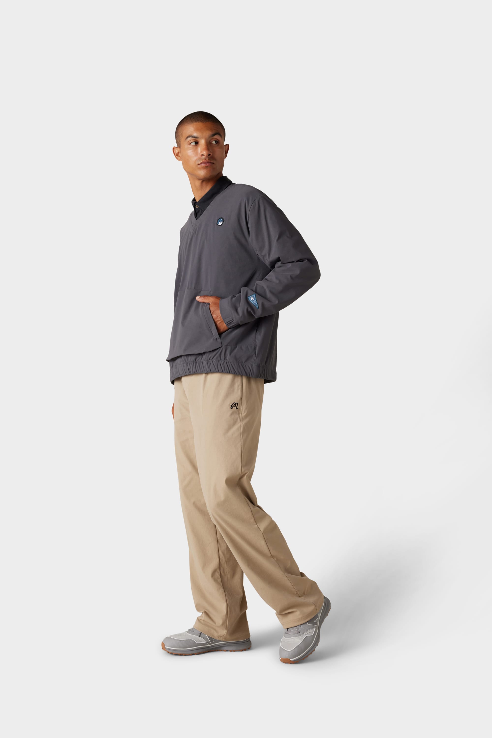 A person stands sideways against a white background, wearing the 686 x Malbon Mens PrimaLoft drirelease® Merino-Lined Pullover Jacket from 686, paired with beige pants and grey sneakers.