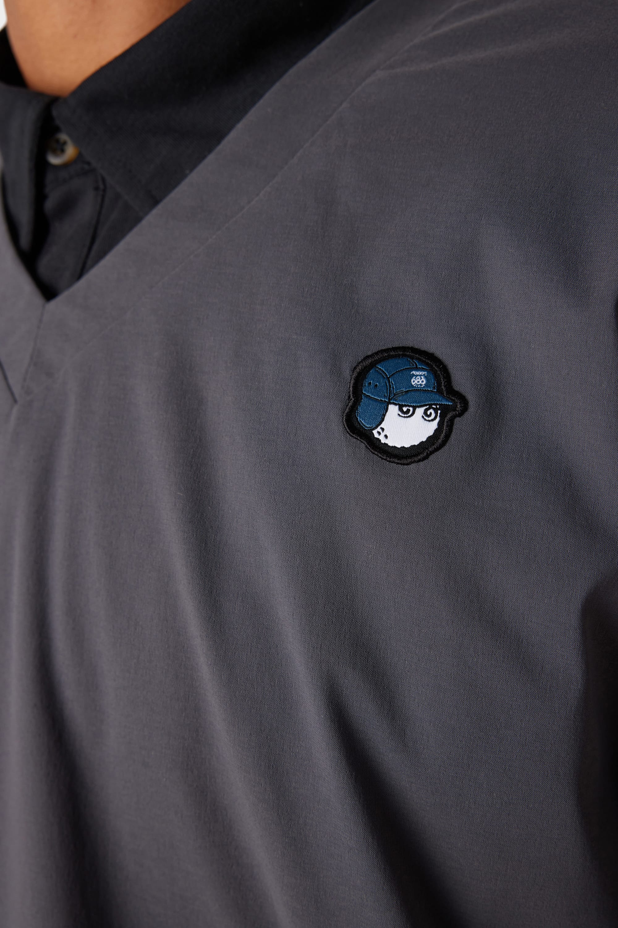 A close-up of the 686 x Malbon Mens PrimaLoft drirelease® Merino-Lined Pullover Jacket in dark grey, featuring a small embroidered baseball with a blue cap on the left chest. The collar and upper part of the 686 jacket are also visible.