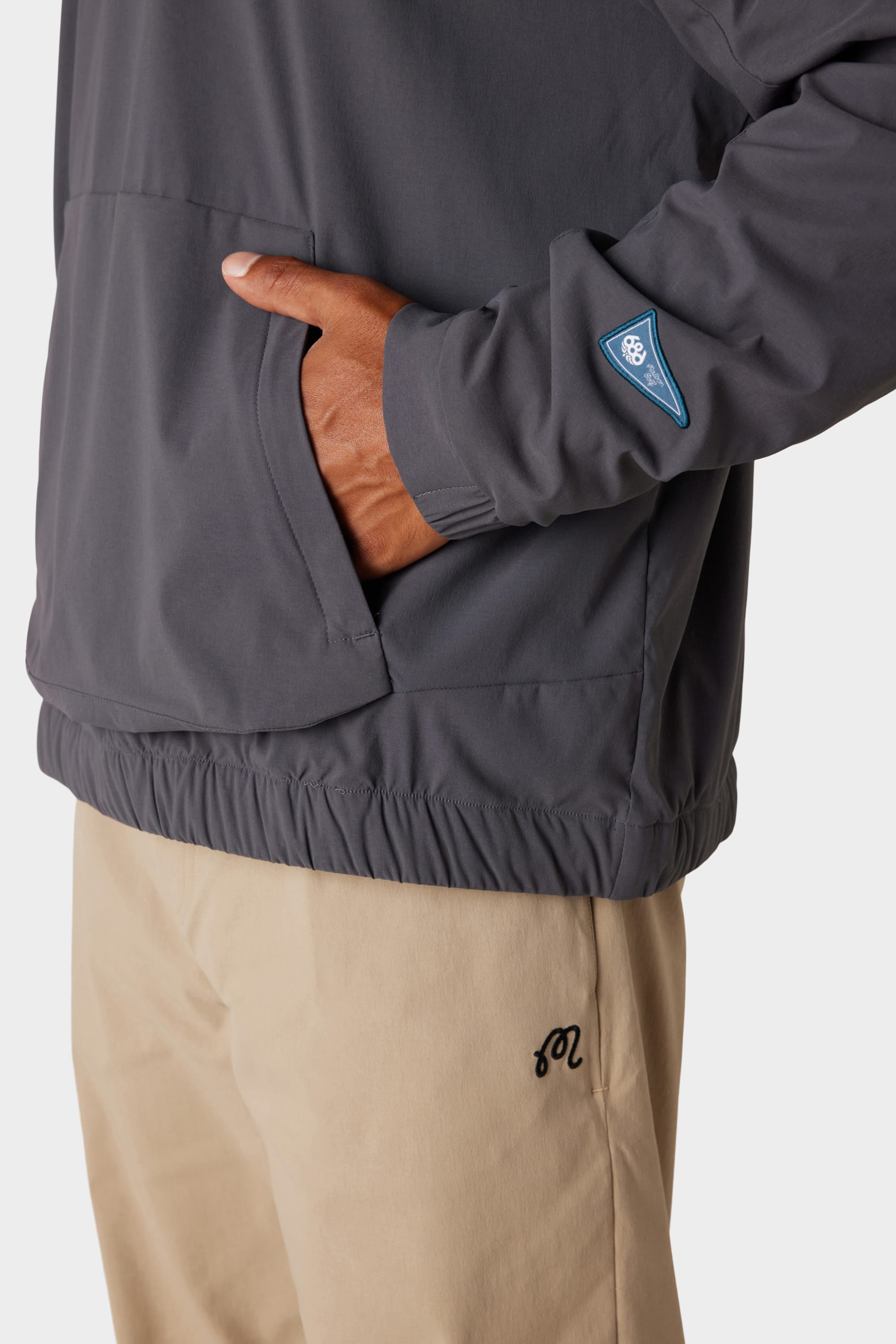 A person is wearing the 686 x Malbon Mens PrimaLoft drirelease® Merino-Lined Pullover Jacket in grey with a blue sleeve patch and tan pants, standing with their right hand in the jackets front pocket. Photo shows only torso and upper legs.