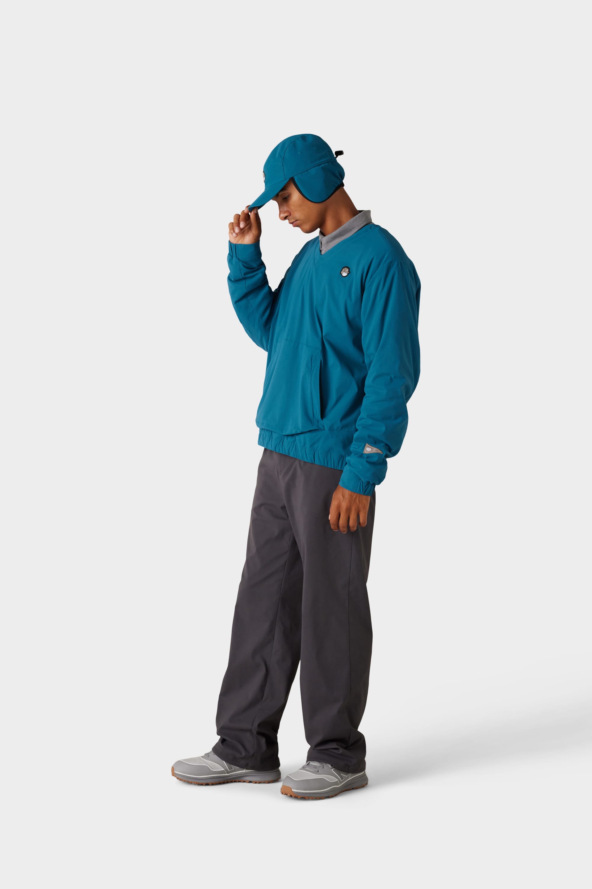 A person wearing the 686 x Malbon Mens PrimaLoft drirelease® Merino-Lined Pullover Jacket in teal, with a matching ear-flap cap, grey pants, and sneakers stands against a white background, holding their caps brim and looking down.
