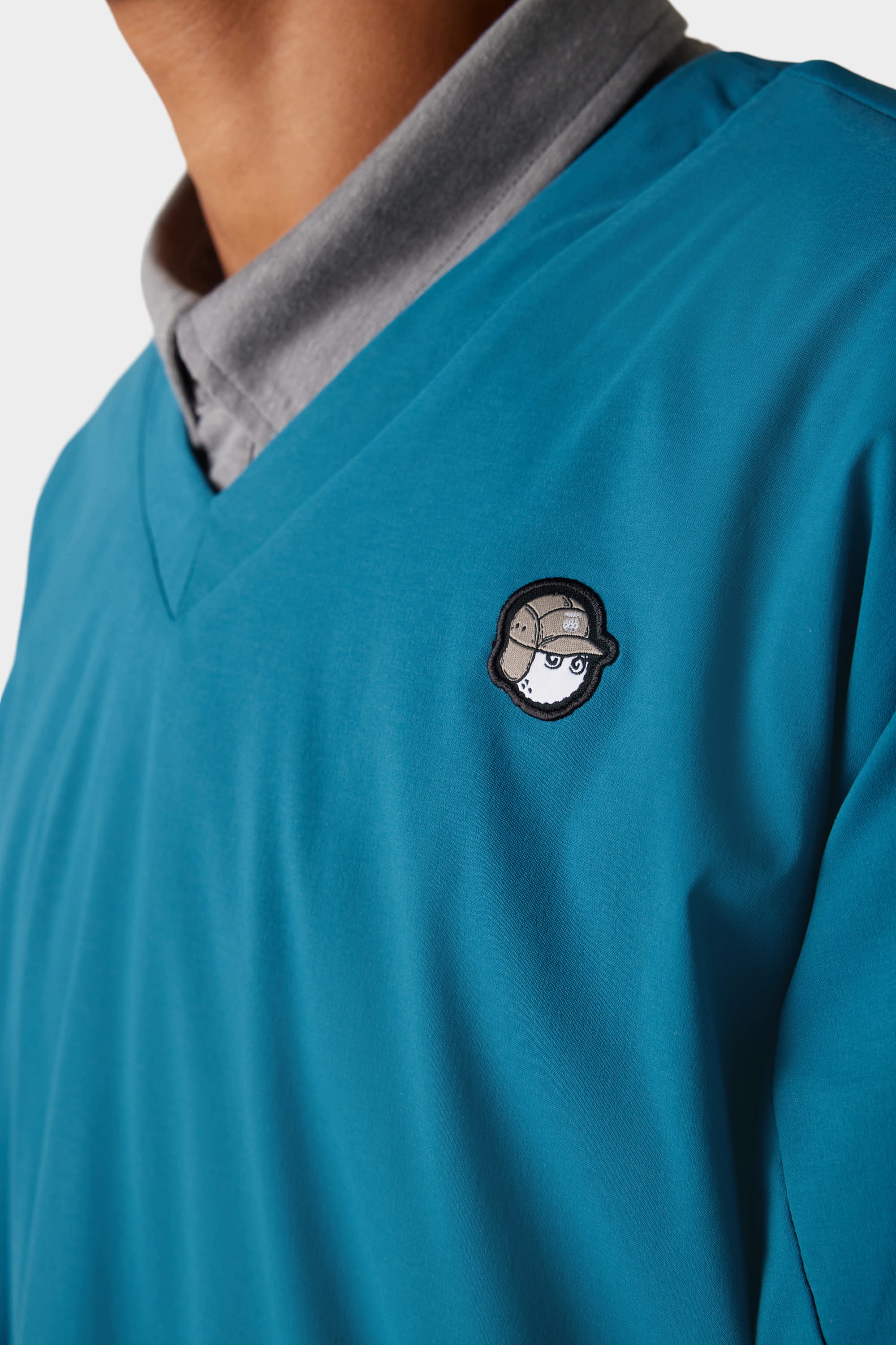 A person wearing a teal 686 x Malbon Mens PrimaLoft drirelease® Merino-Lined Pullover Jacket over a grey collared shirt. The jacket has a small embroidered cartoon face with a helmet on the left chest.