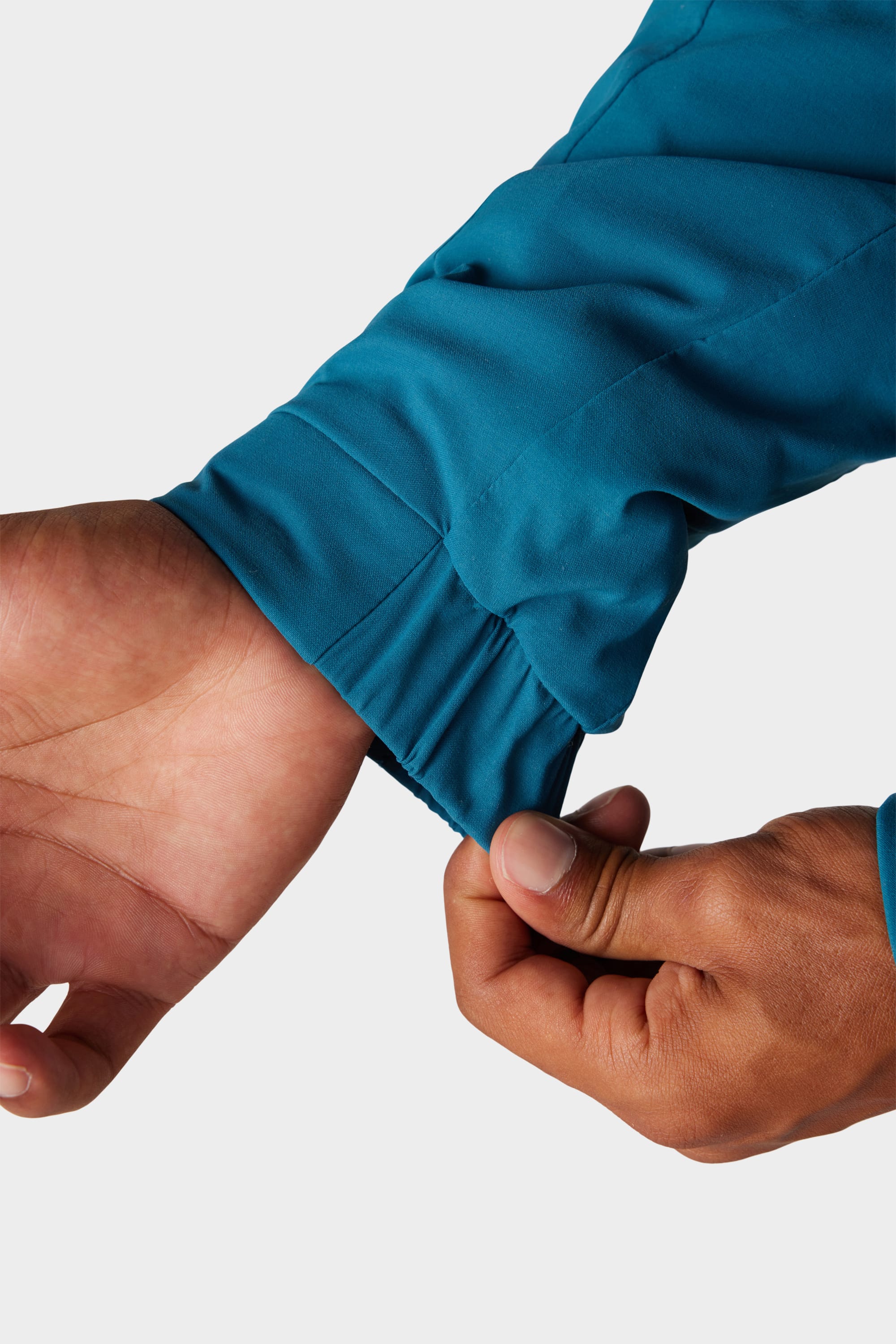 A hand adjusts the elastic cuff of a blue 686 x Malbon Mens PrimaLoft drirelease® Merino-Lined Pullover Jacket, highlighting its stretchiness against a plain, light background.