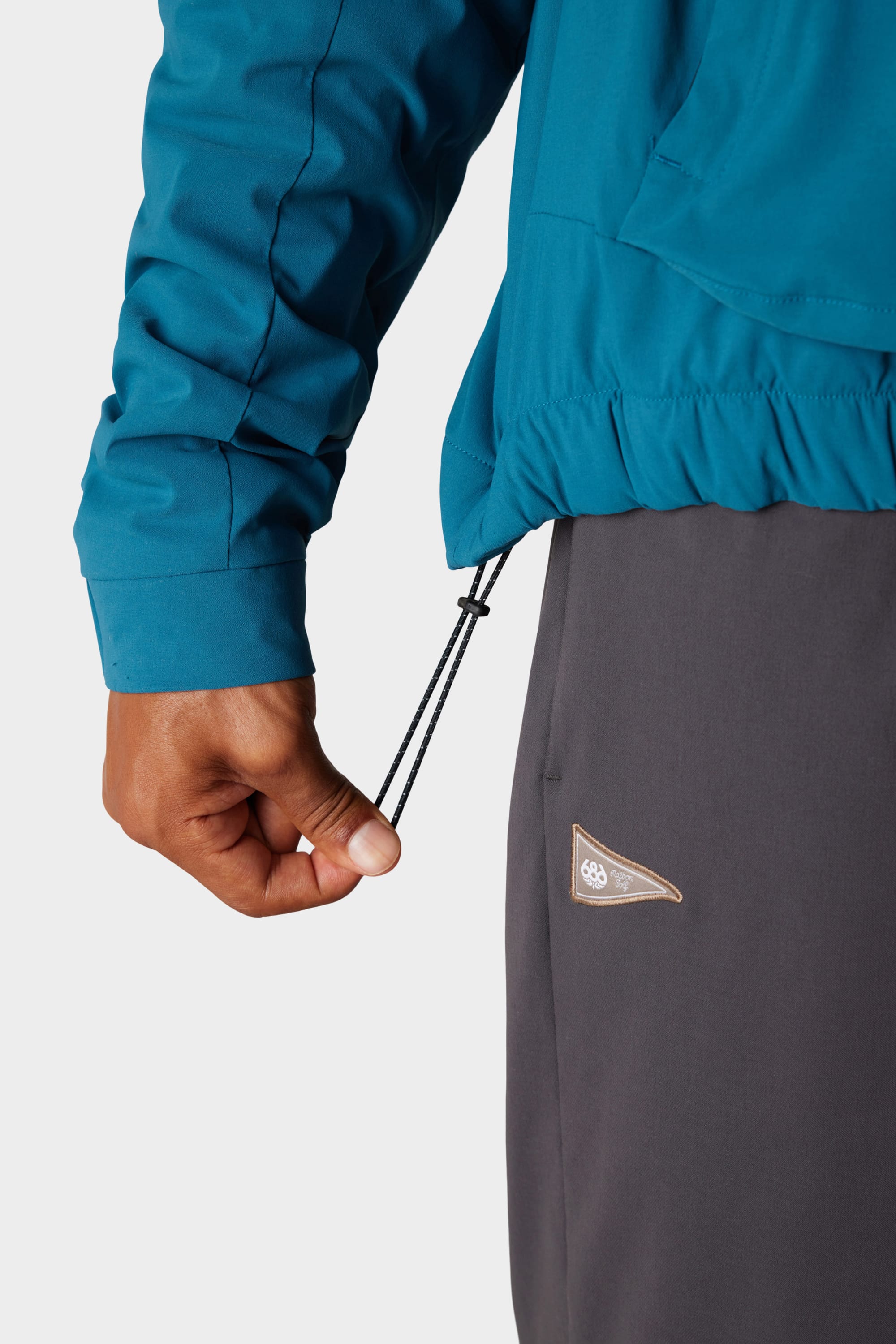 A person adjusts the hem drawcord on a teal 686 x Malbon Mens PrimaLoft drirelease® Merino-Lined Pullover Jacket, paired with dark grey pants featuring a small triangular logo patch near the pocket.