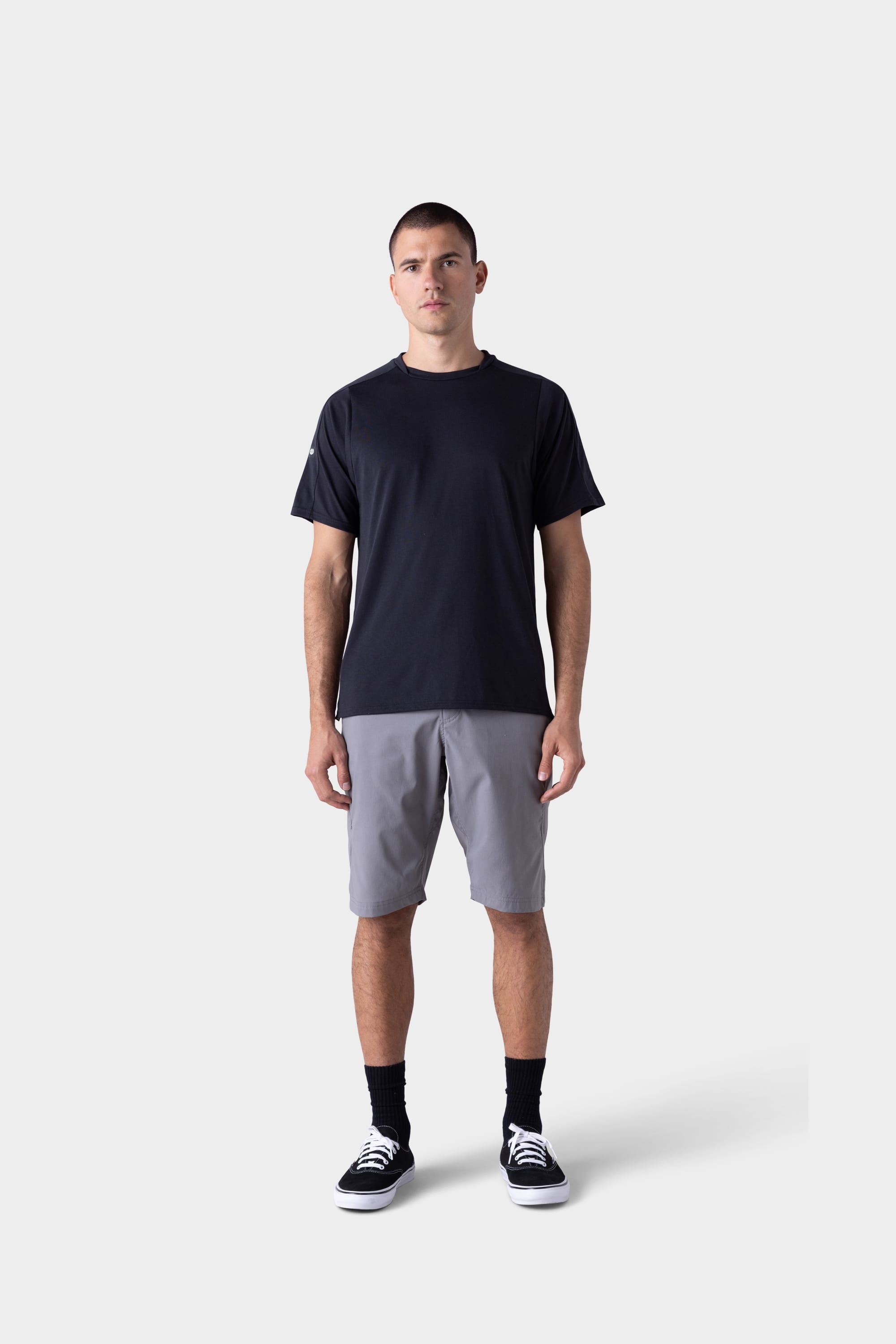 A man stands facing forward against a plain light grey background, wearing the 686 Mens Rival Short Sleeve Bike Jersey in black, paired with grey shorts, black socks, and black sneakers with white soles.