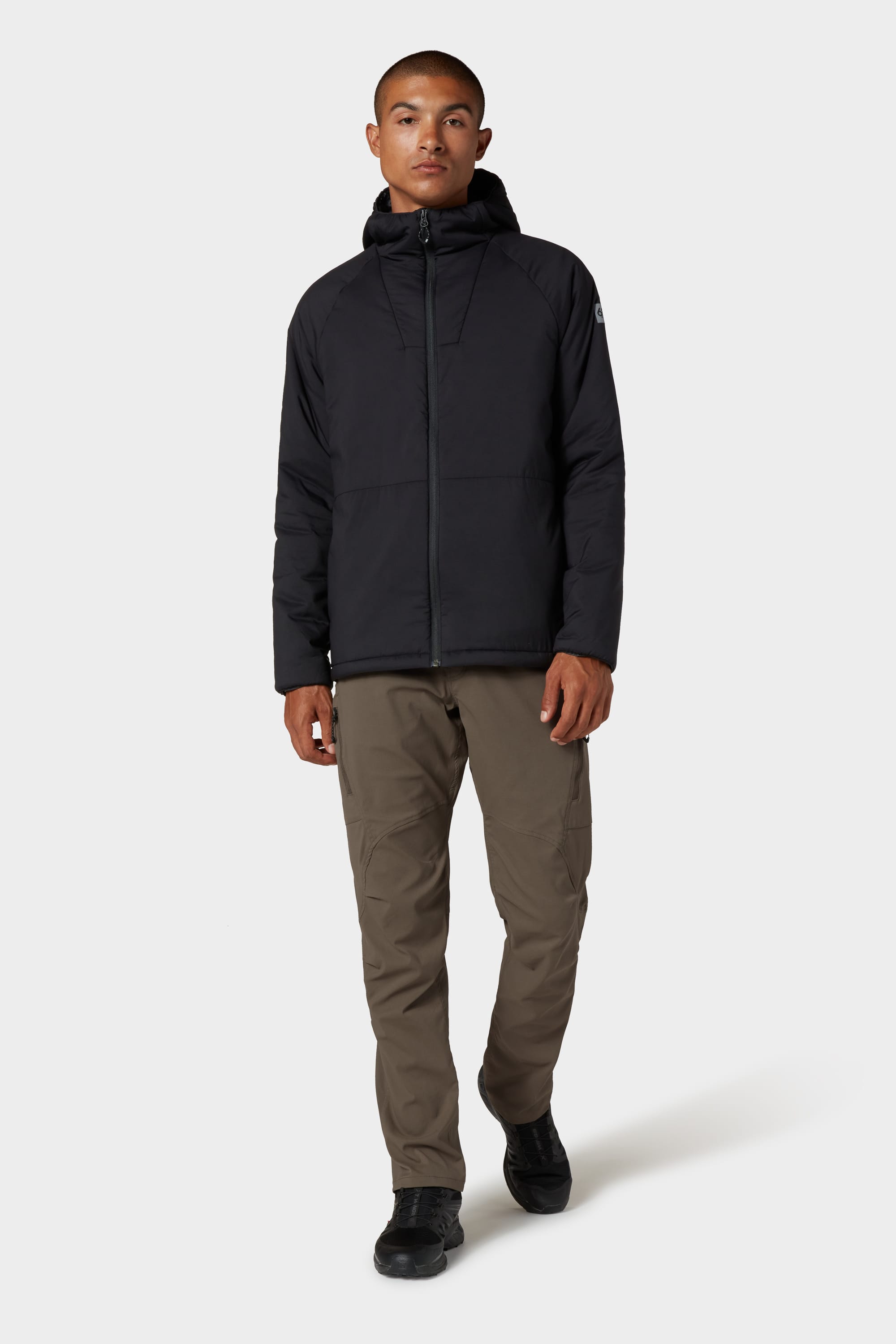 A man stands against a plain background, facing forward with a neutral expression, wearing the 686 Mens PrimaLoft® Source Jacket in black by 686, paired with light brown pants and black shoes.