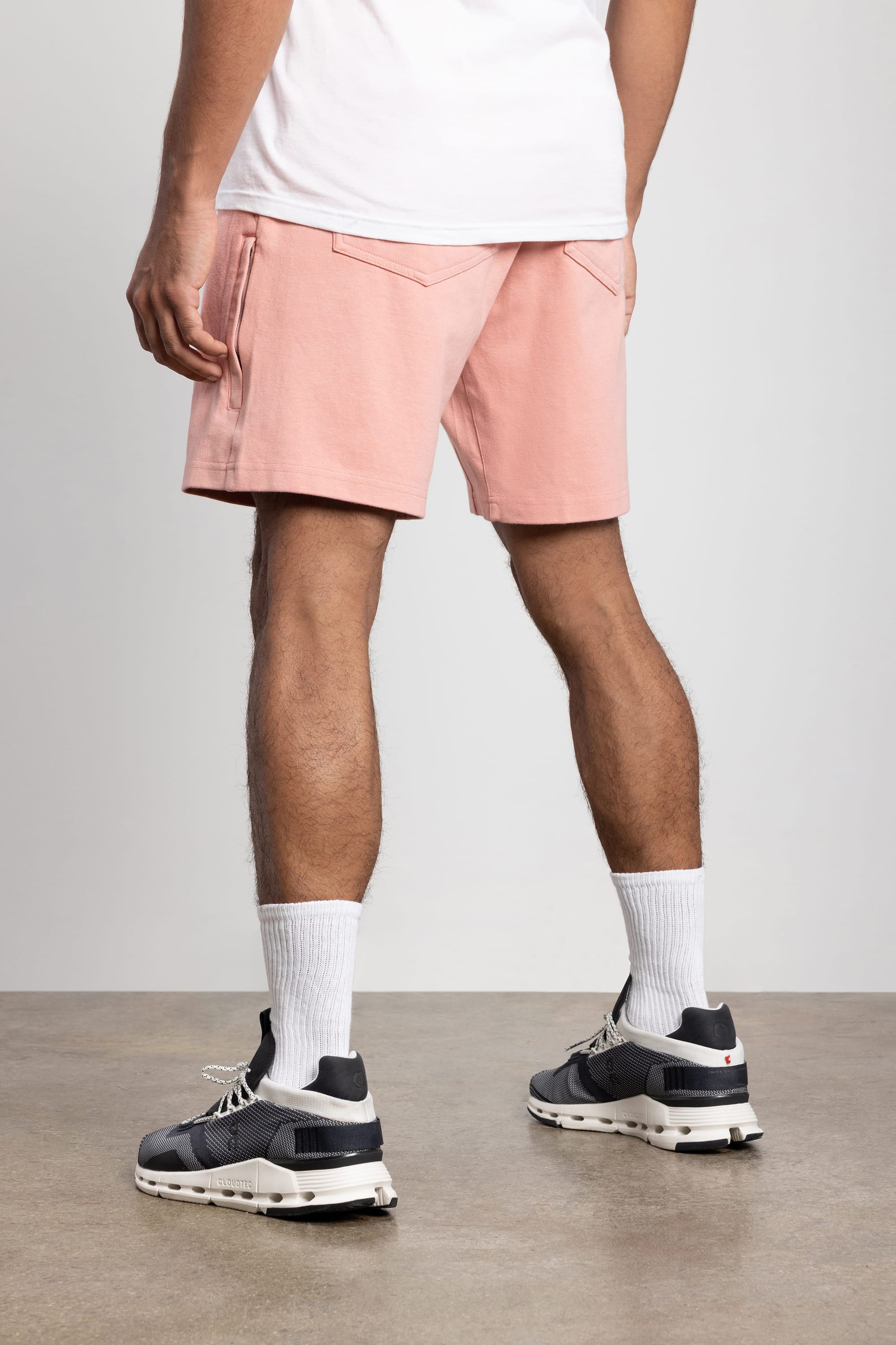A person stands indoors on a concrete floor, wearing a white shirt, white crew socks, black athletic shoes, and 686 Mens Everywhere Double Knit Short in light pink. The photo is taken from behind, showing only the lower half of the body.