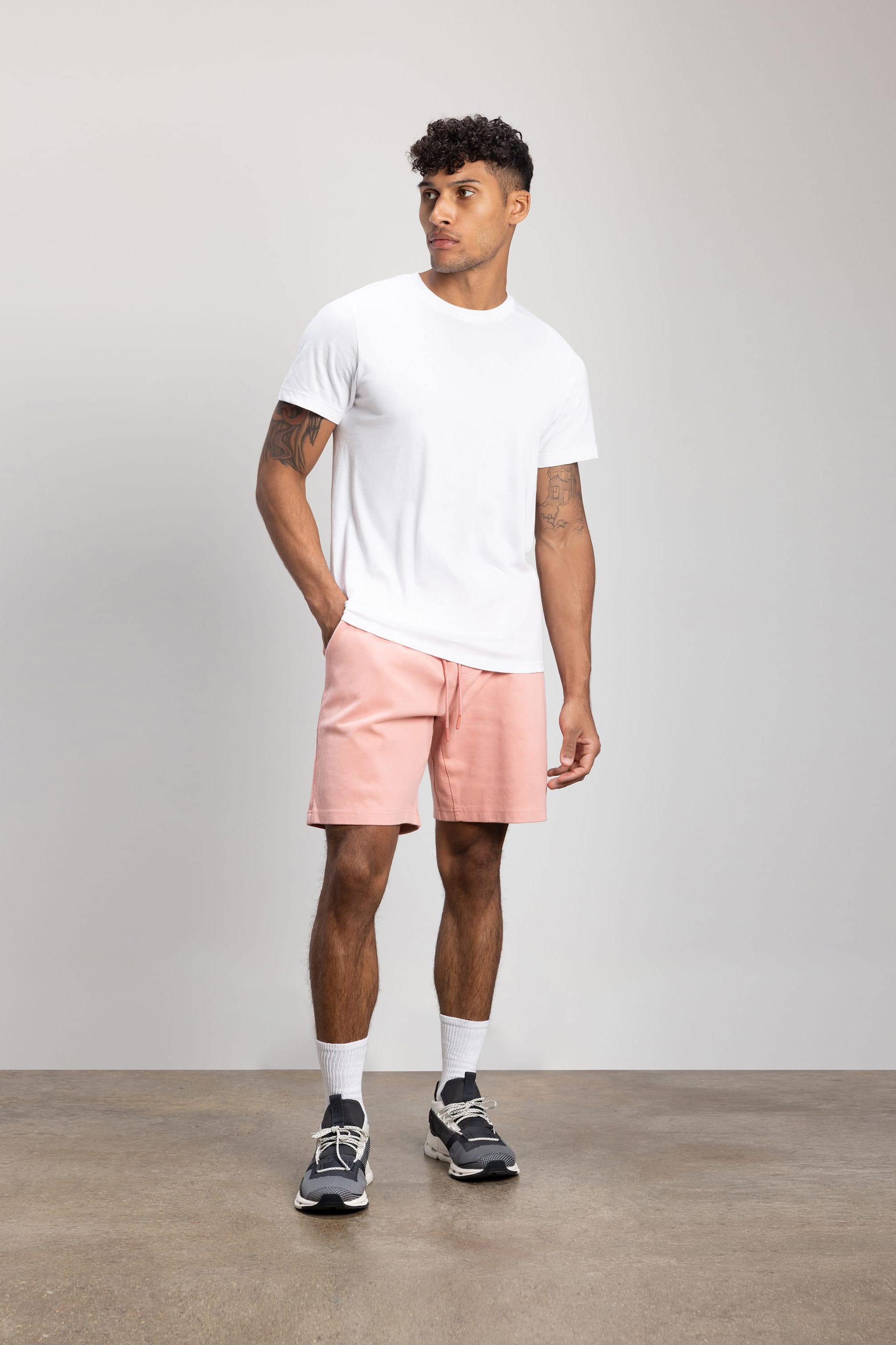 A man stands against a plain background wearing a white t-shirt, 686 Mens Everywhere Double Knit Short in light pink, white socks, and black sneakers. His left hand is in his pocket as he looks slightly to his right.