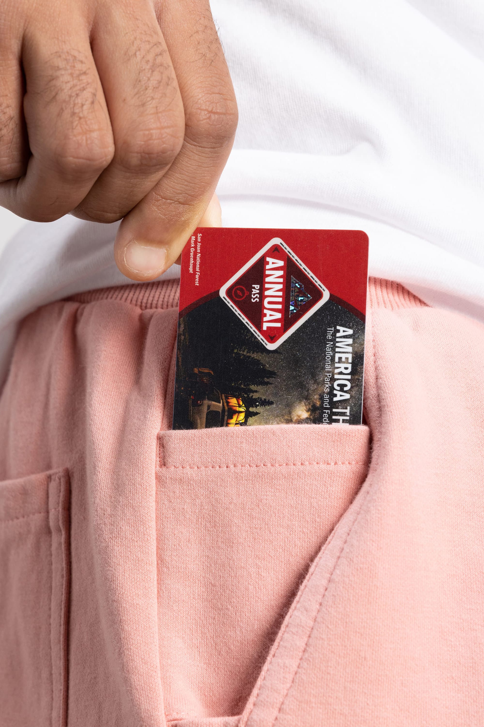 A hand is seen putting a red America the Beautiful annual pass into the front pocket of pink 686 Mens Everywhere Double Knit Shorts by 686. The person is wearing a white shirt.