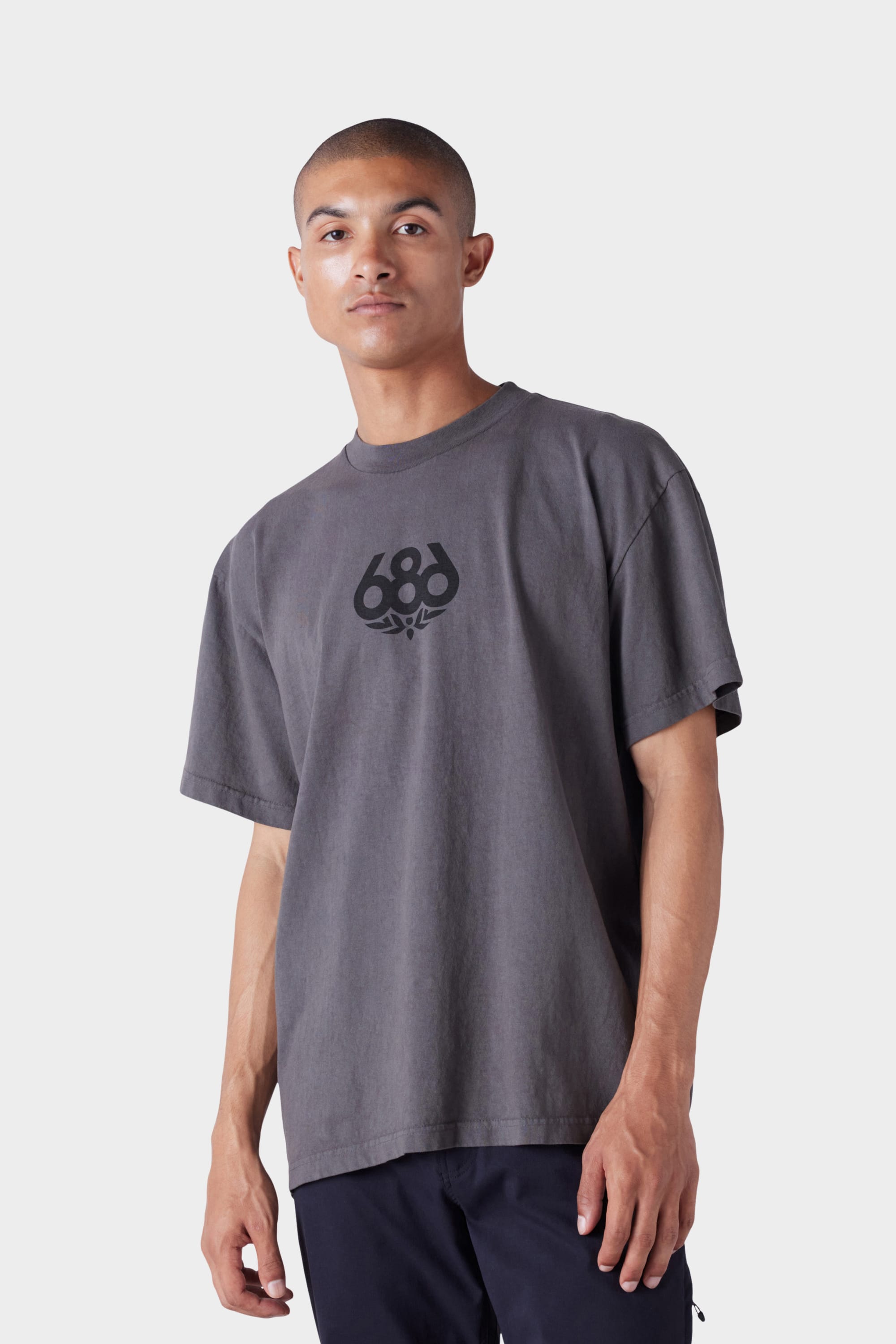 A man with short hair and a neutral expression wears the 686 Mens Wreath Short Sleeve Tee—a loose-fitting, faded grey t-shirt with a black 686 logo on the chest—against a plain light grey background.