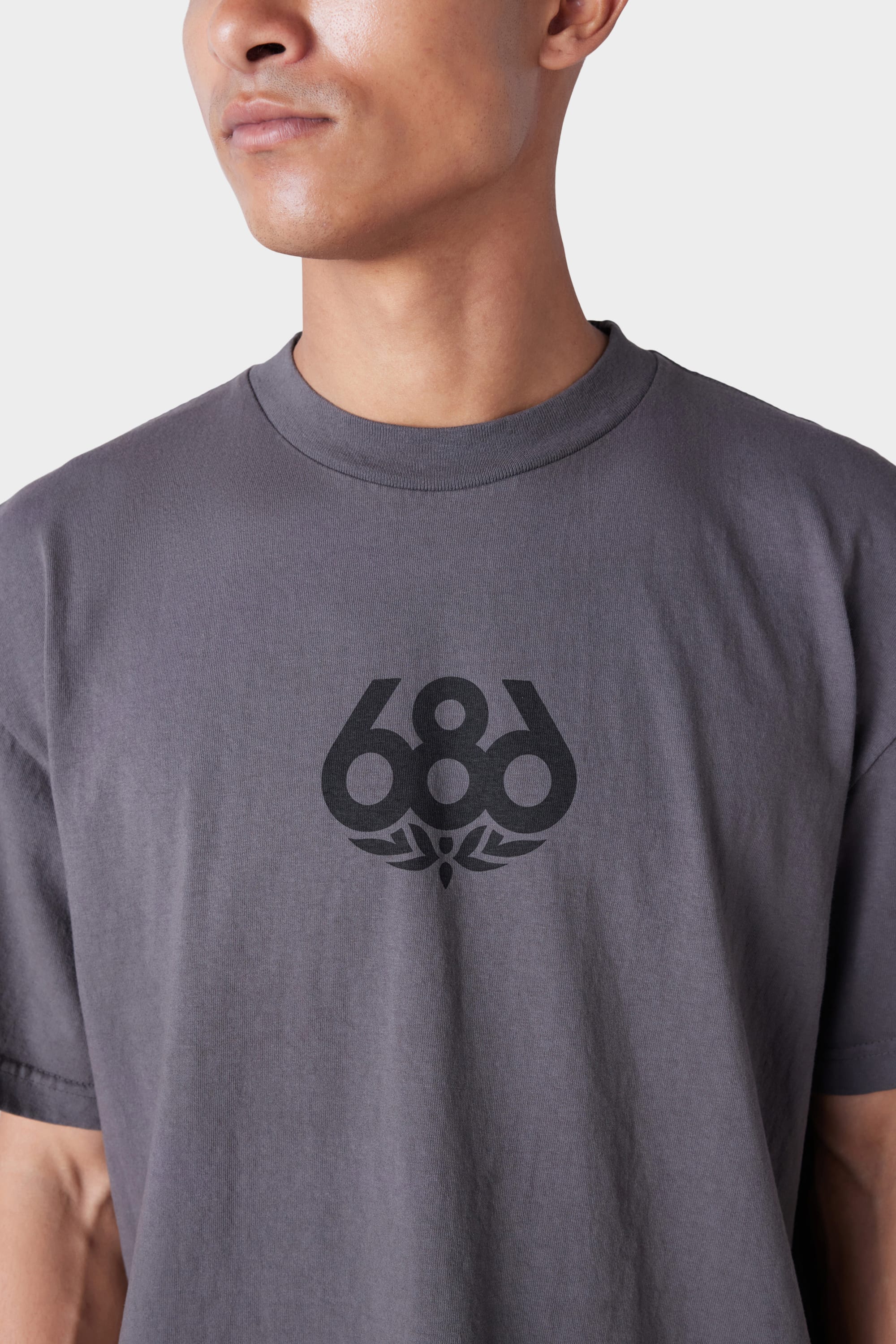 A man is shown from the shoulders up against a plain background, wearing the 686 Mens Wreath Short Sleeve Tee—a grey crew-neck T-shirt featuring the black 686 logo with a stylized winged design beneath it.