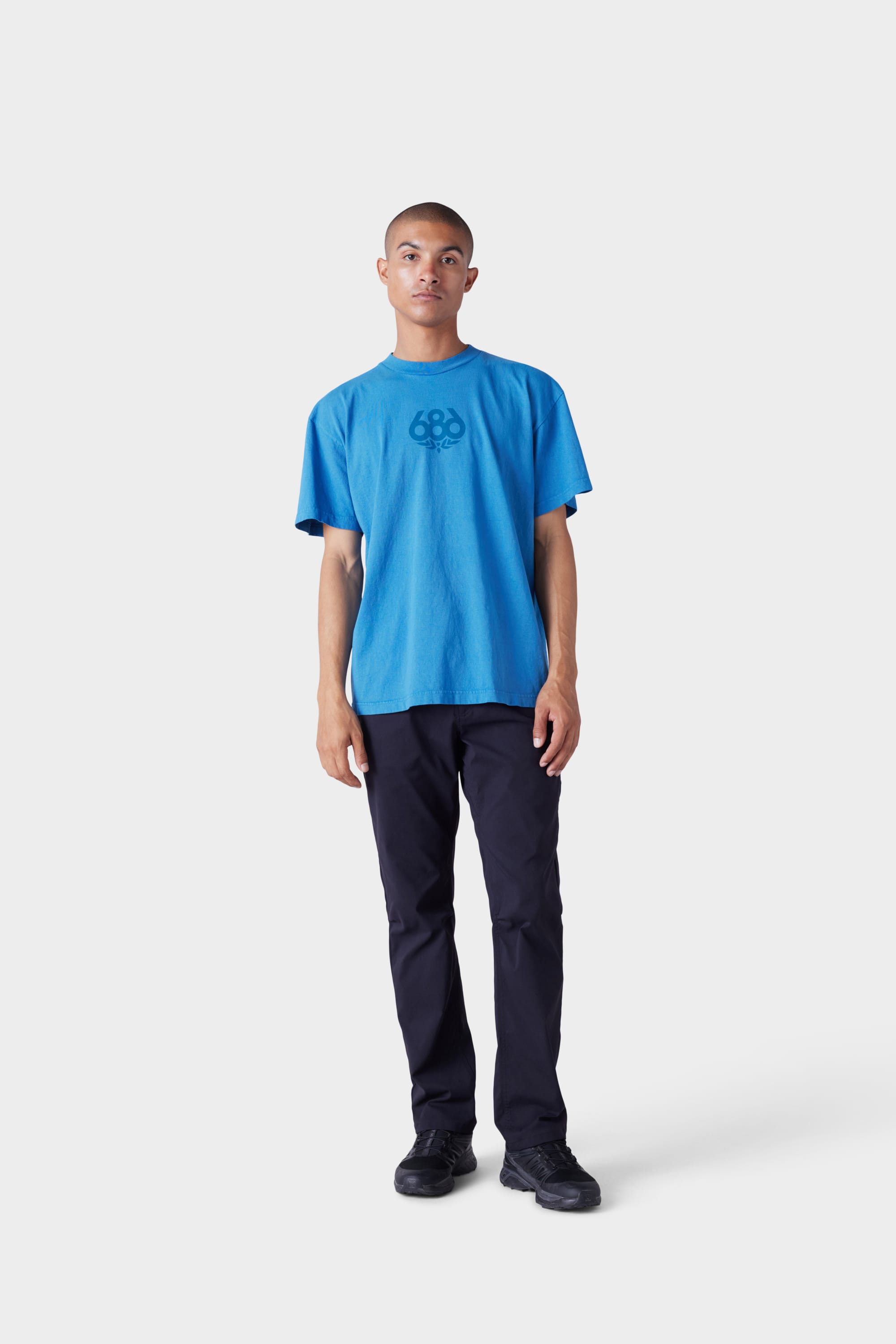 A person stands against a plain white background, wearing the 686 Mens Wreath Short Sleeve Tee by 686, paired with black pants and black shoes.