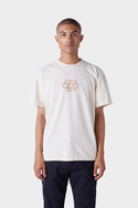 A person faces forward against a plain light grey background, wearing the 686 Mens Wreath Short Sleeve Tee in light beige with a central 686 graphic, paired with dark pants.