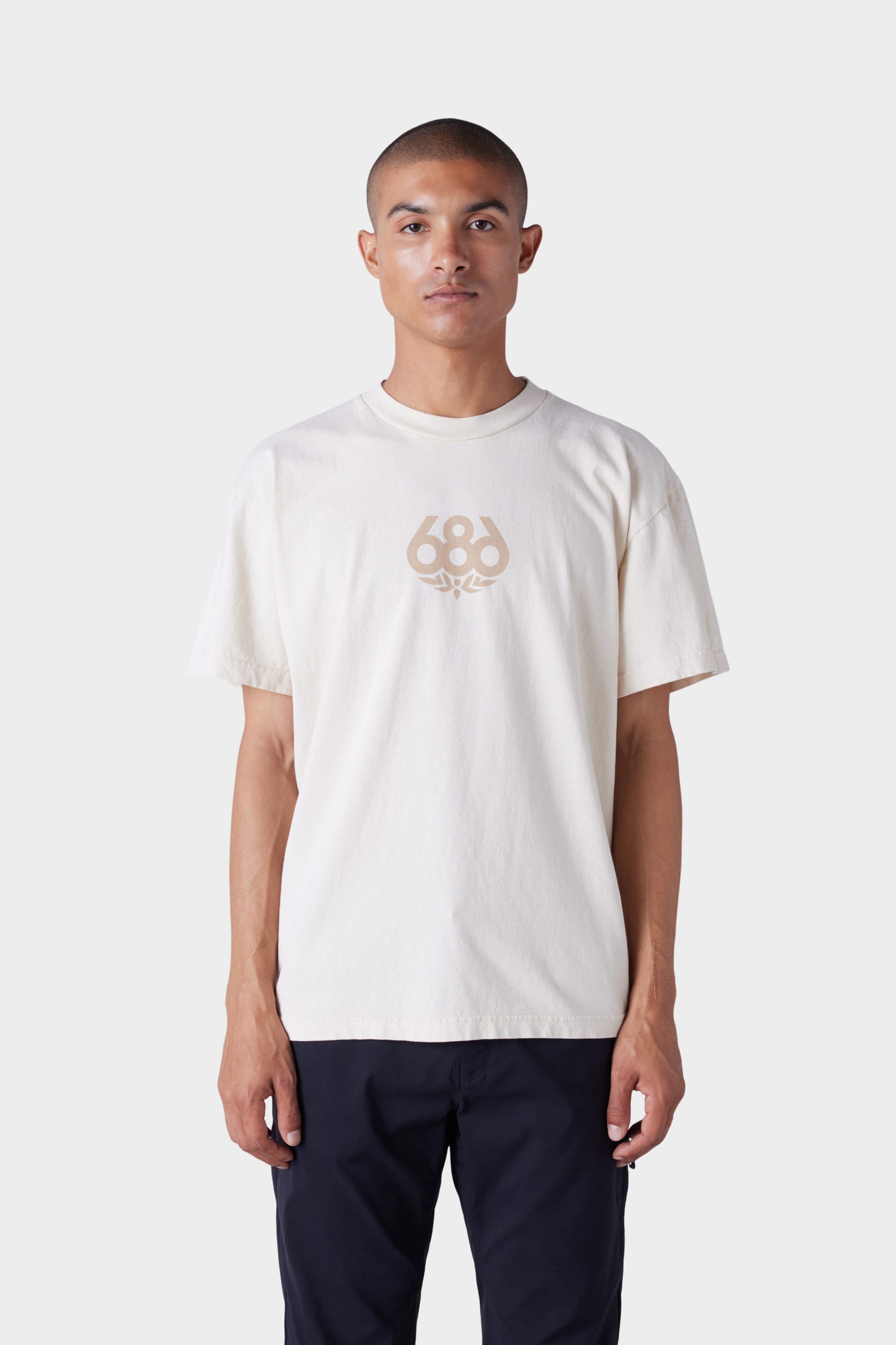 A person faces forward against a plain light grey background, wearing the 686 Mens Wreath Short Sleeve Tee in light beige with a central 686 graphic, paired with dark pants.
