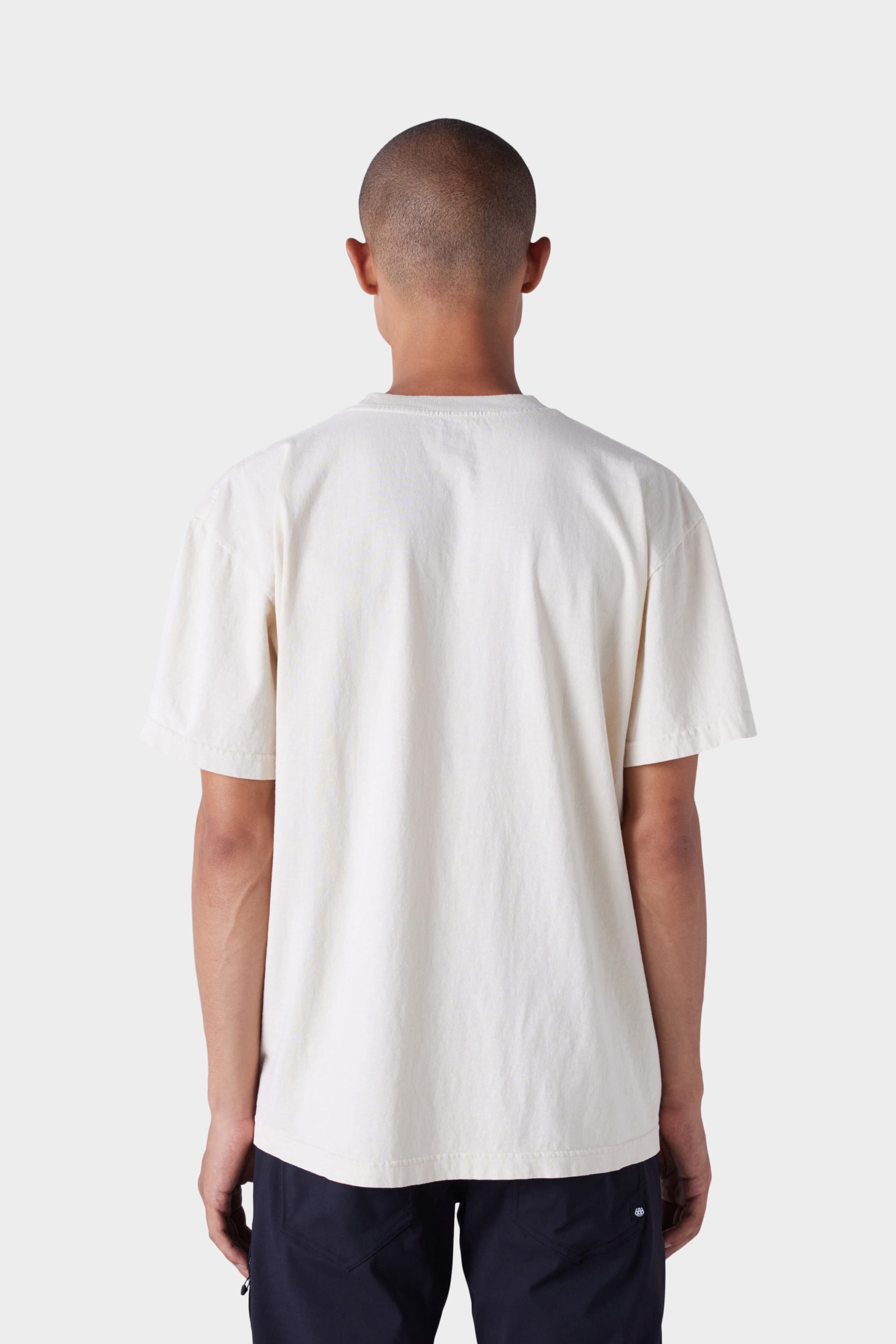 A person with a shaved head stands with their back to the camera, wearing the 686 Mens Wreath Short Sleeve Tee by 686 and dark pants against a light grey background.