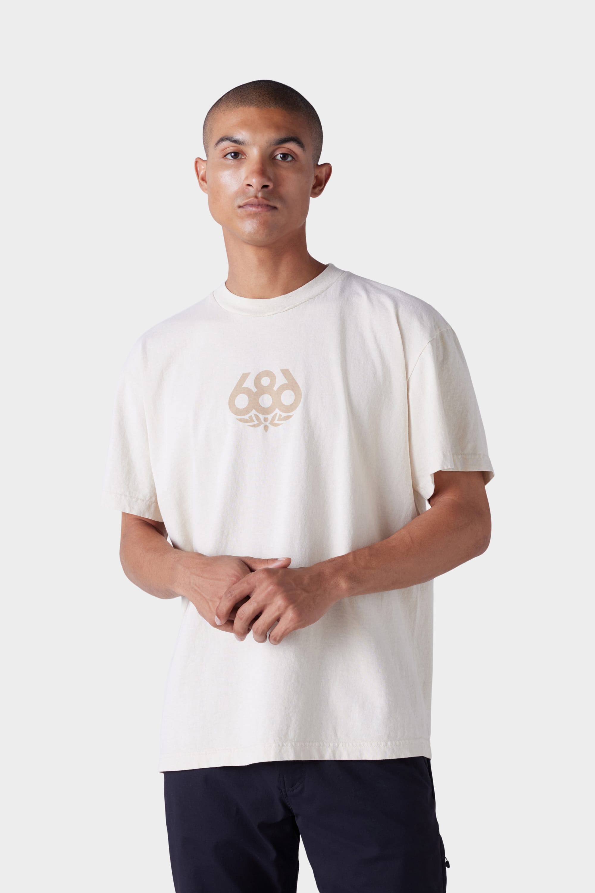 A man stands facing forward against a light grey background, wearing the 686 Mens Wreath Short Sleeve Tee in a light color with the 686 logo on the front and his hands loosely clasped.