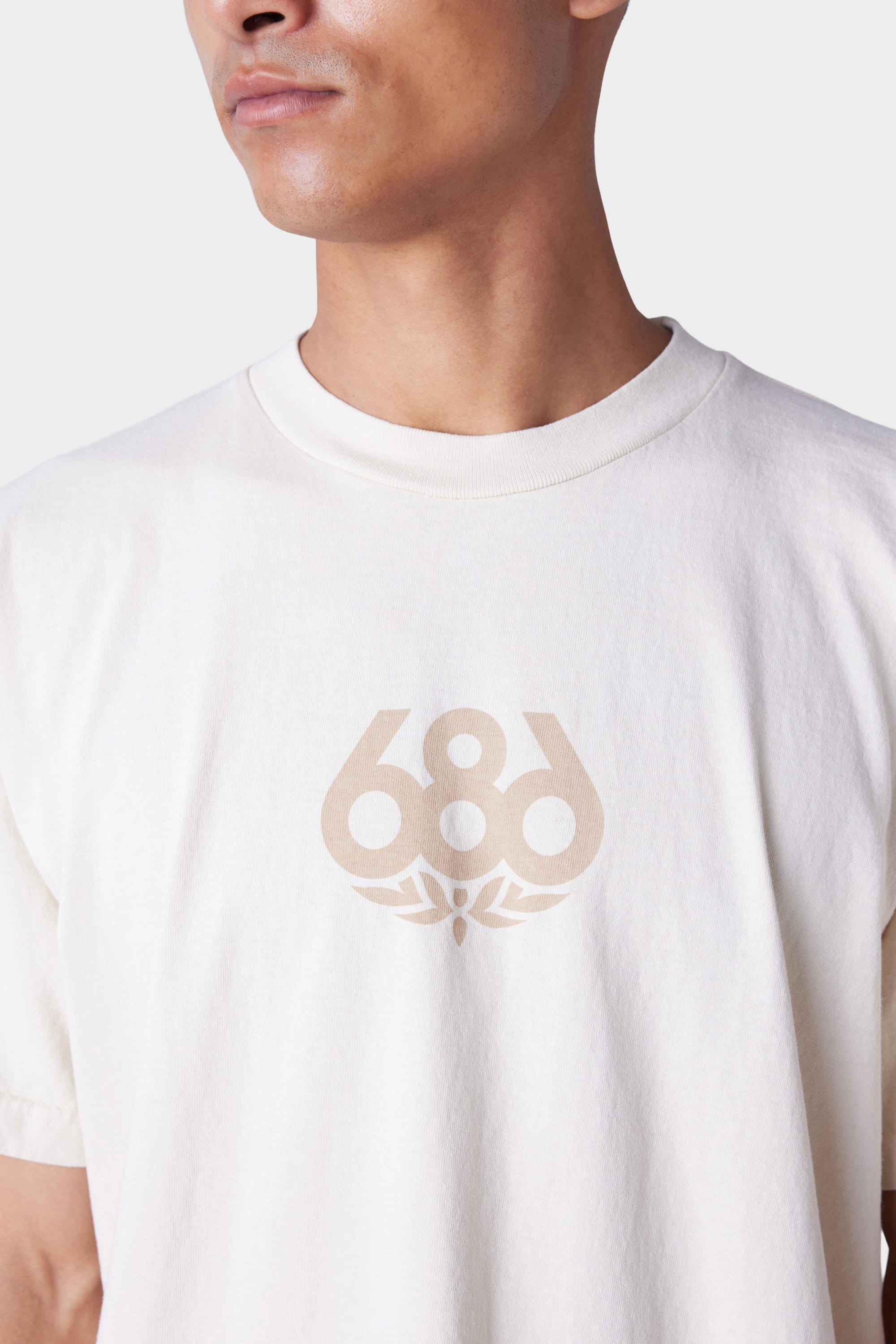 A person wearing the 686 Mens Wreath Short Sleeve Tee, an off-white T-shirt featuring the beige 686 logo with a small graphic beneath it on the chest. Only their lower face and upper torso are visible.