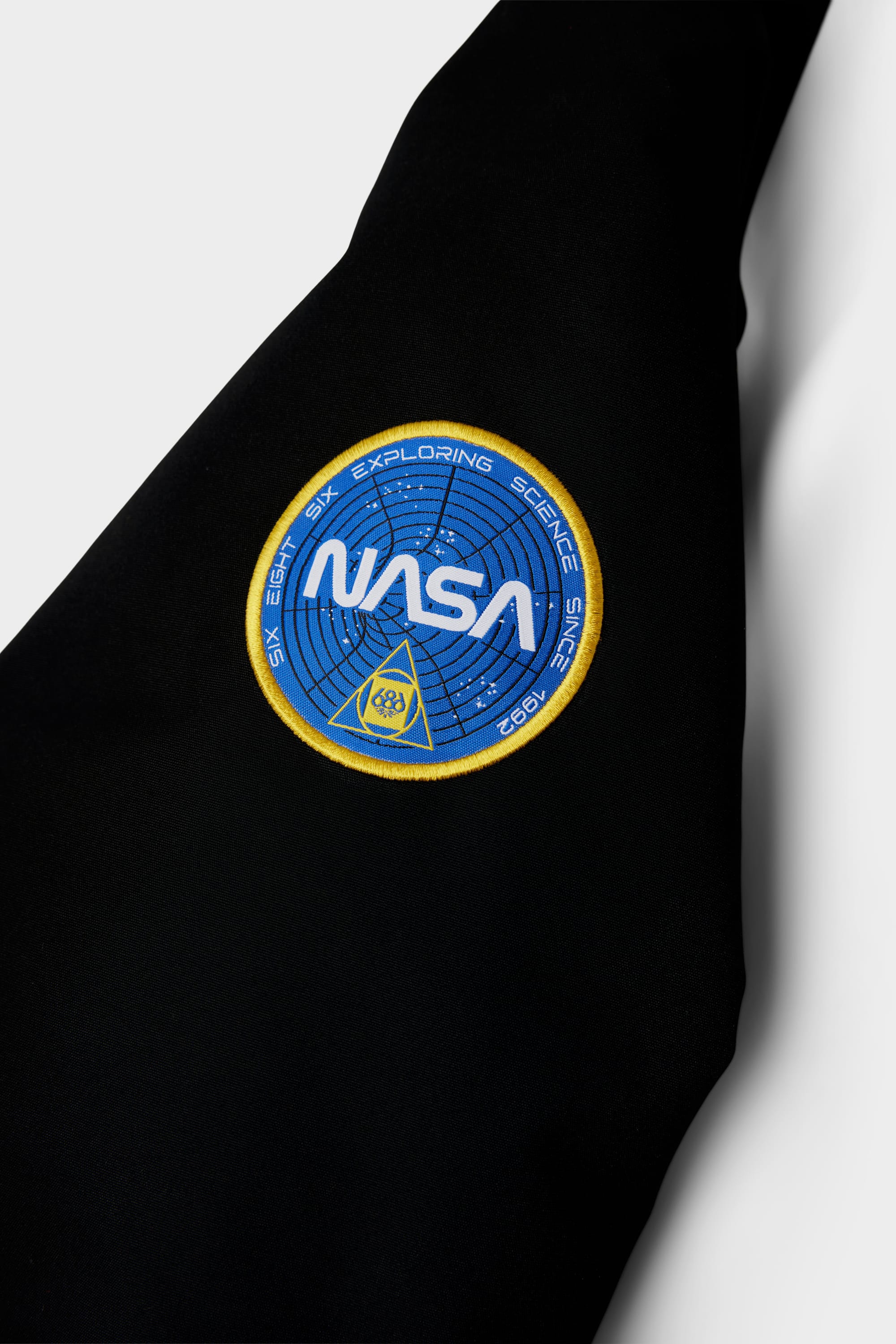 A close-up of the 686 Youth Exploration Insulated Jacket by 686, showing a black fabric with a NASA patch featuring a blue circle, white NASA text, and Exploring Science Since 1958 around the edge.