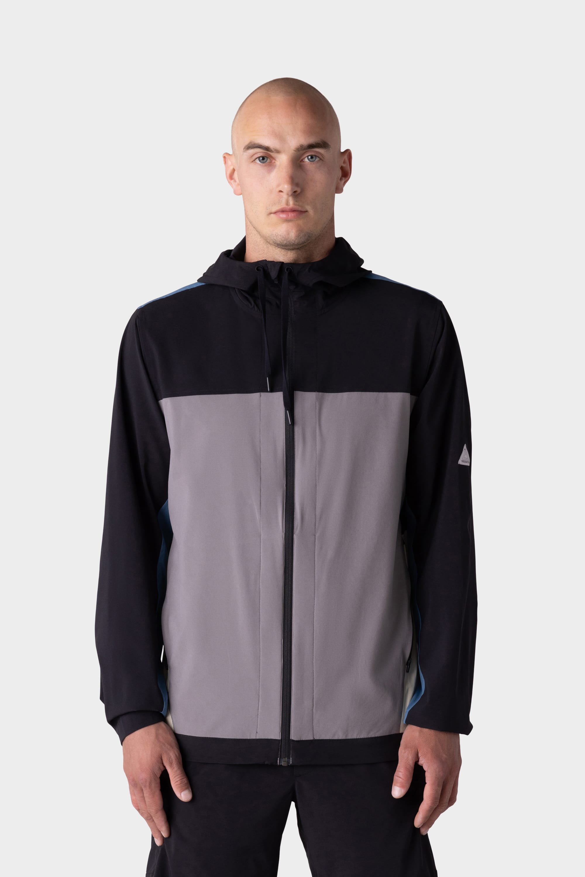 A man with a shaved head wears the 686 Mens ATP Stretch Wind Jacket by 686, featuring black sleeves and a gray front panel, standing against a light gray background.