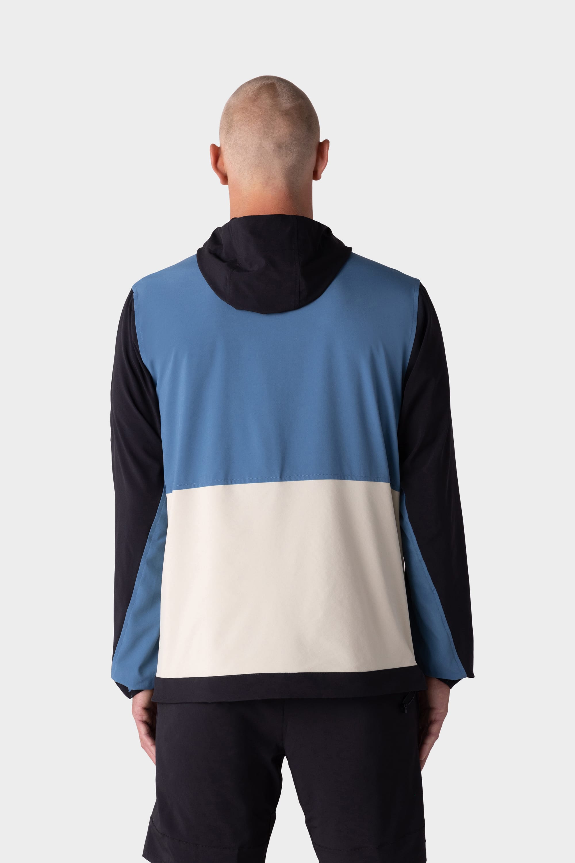 A person with a shaved head is seen from behind wearing the 686 Mens ATP Stretch Wind Jacket by 686, featuring blue, white, and black color-block panels, paired with black shorts against a plain light gray background.