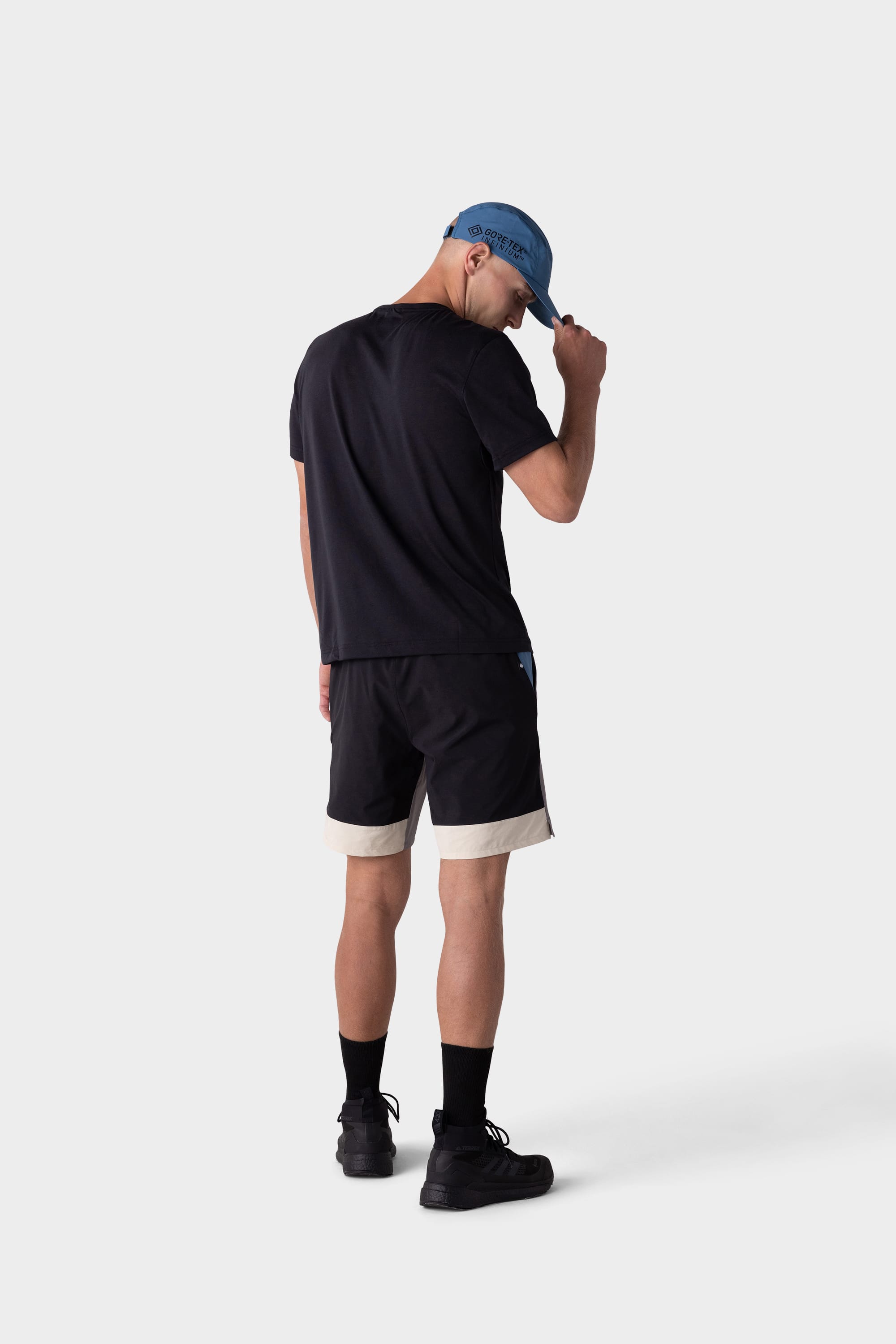 A person stands facing away, wearing a black t-shirt, 686 Mens ATP Stretch Performance Shorts by 686 in black and beige, black socks and shoes, and a light blue cap, adjusting the cap with one hand against a plain light gray background.