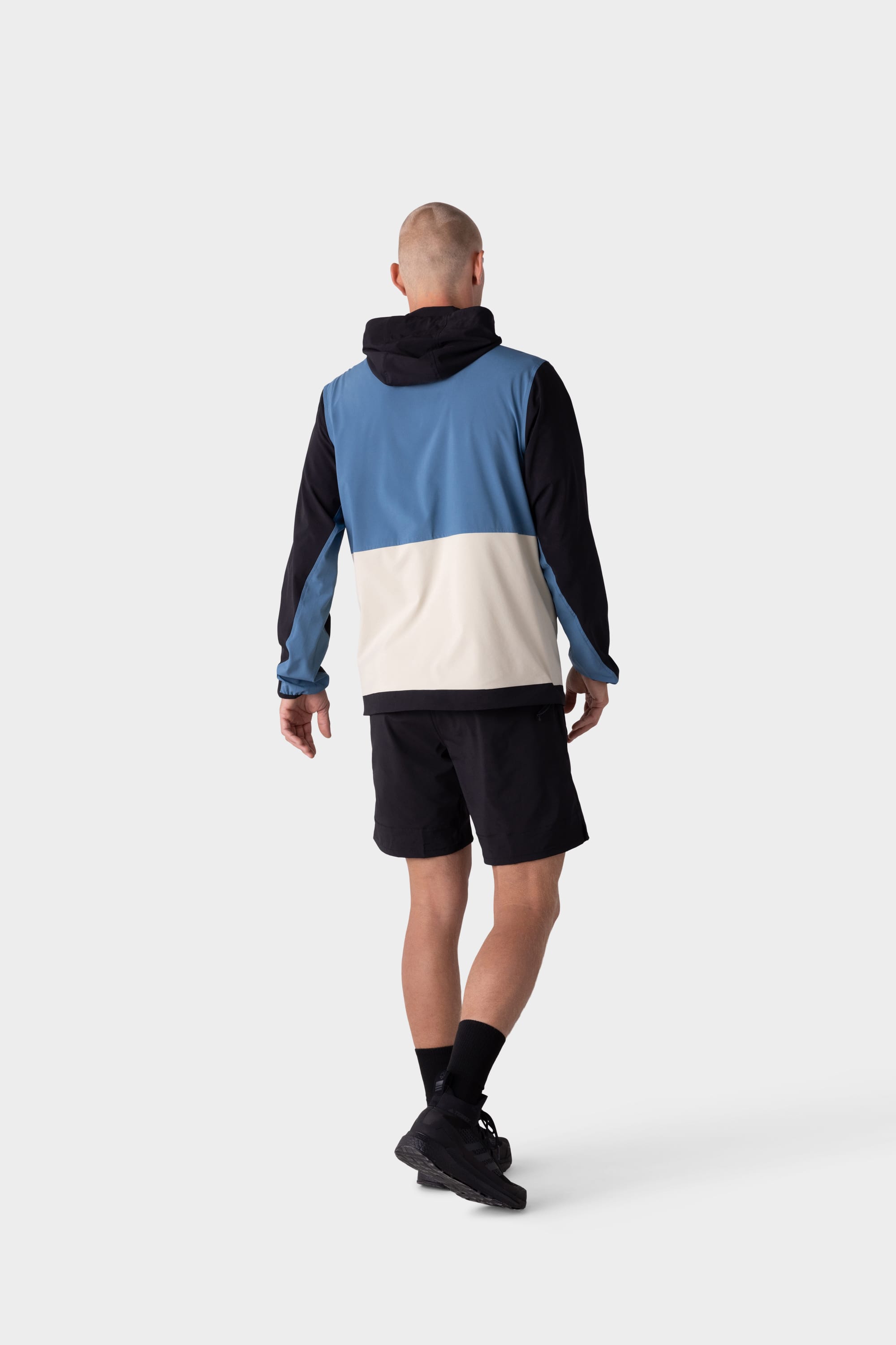 A man stands facing away in a 686 Mens ATP Stretch Wind Jacket by 686, paired with black shorts, black socks, and black sneakers, against a plain light gray background.