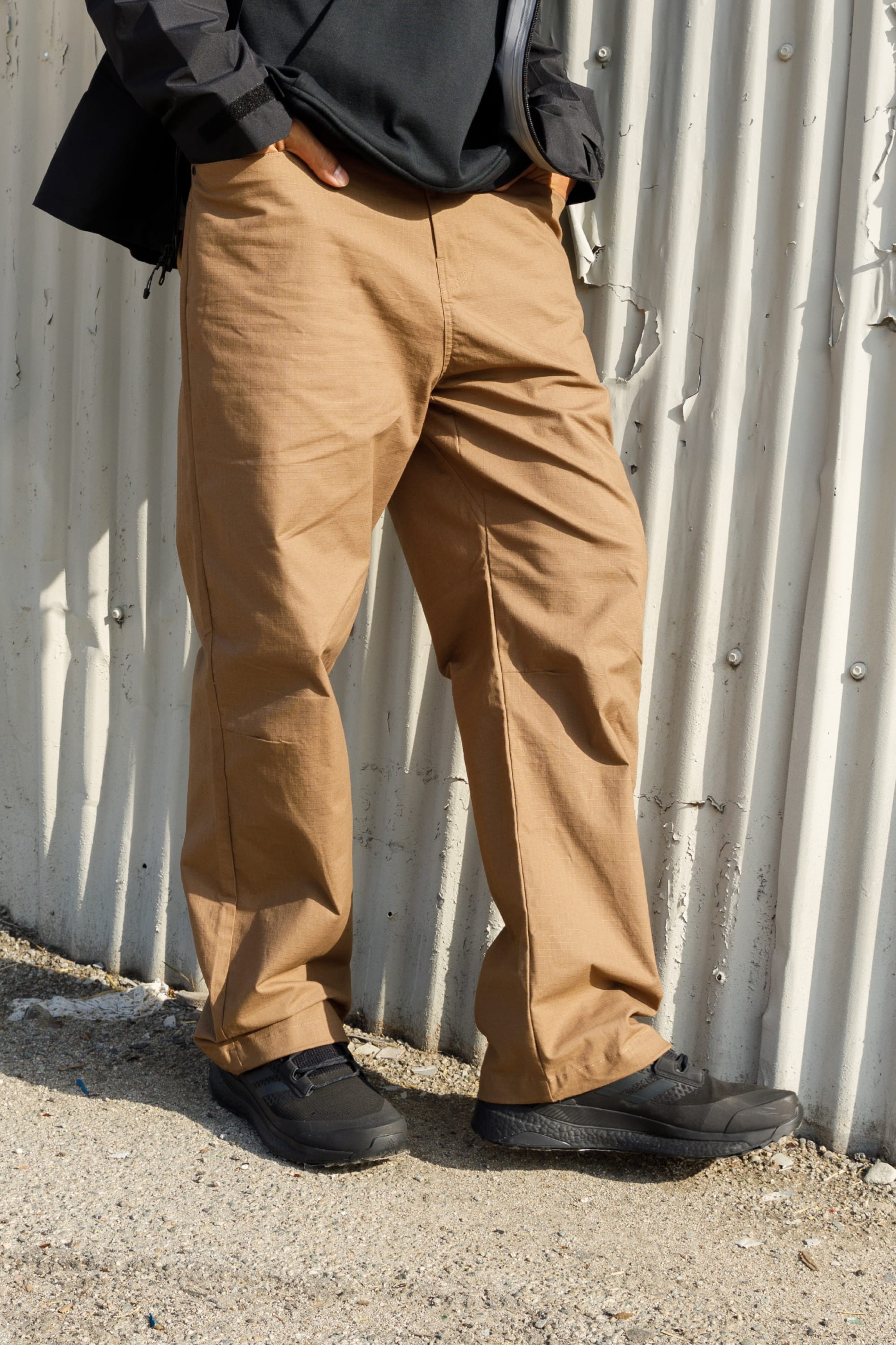 A person stands outdoors by a corrugated metal wall, hands in pockets, wearing only visible lower-body apparel: the 686 Mens Cruiser Pant - Wide Fit in tan.