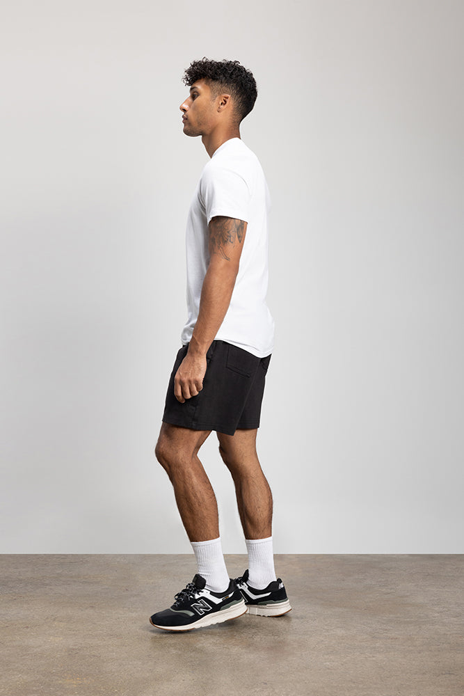A man with short curly hair stands sideways against a light gray background, wearing a white t-shirt, 686 Mens Everywhere Double Knit Short by 686, white socks, and black sneakers.