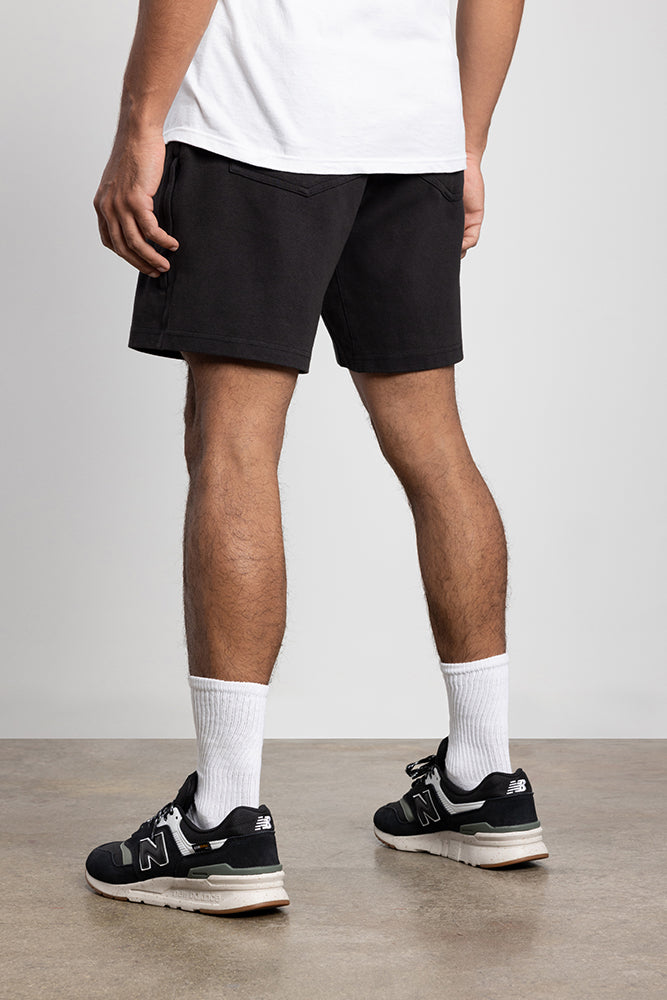 A person stands on a concrete floor wearing a white T-shirt, 686 Mens Everywhere Double Knit Shorts, white crew socks, and black New Balance sneakers. The photo is taken from behind, showing their legs and lower back.