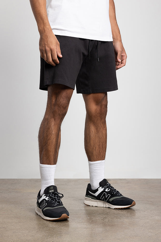 A person wearing a white T-shirt, 686 Mens Everywhere Double Knit Short in black, white crew socks, and black New Balance sneakers stands on a smooth concrete floor against a plain light gray wall.