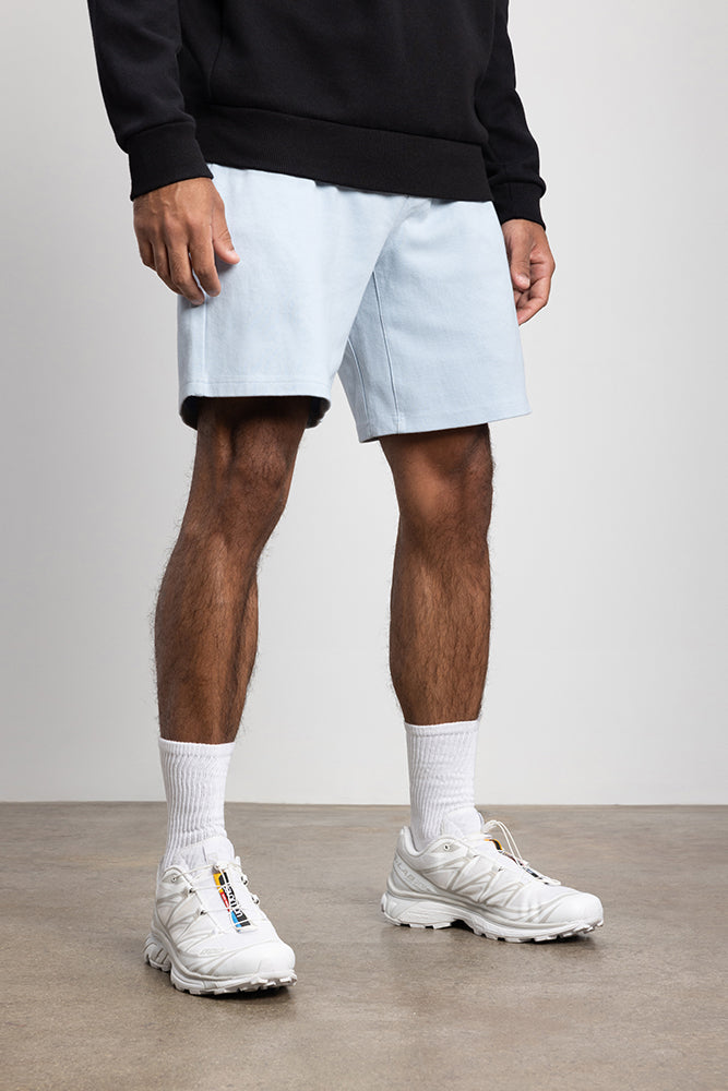 A person wearing a black sweatshirt, 686 Mens Everywhere Double Knit Short by 686 in light blue, white crew socks, and white sneakers stands on a concrete floor against a plain, light-colored background.
