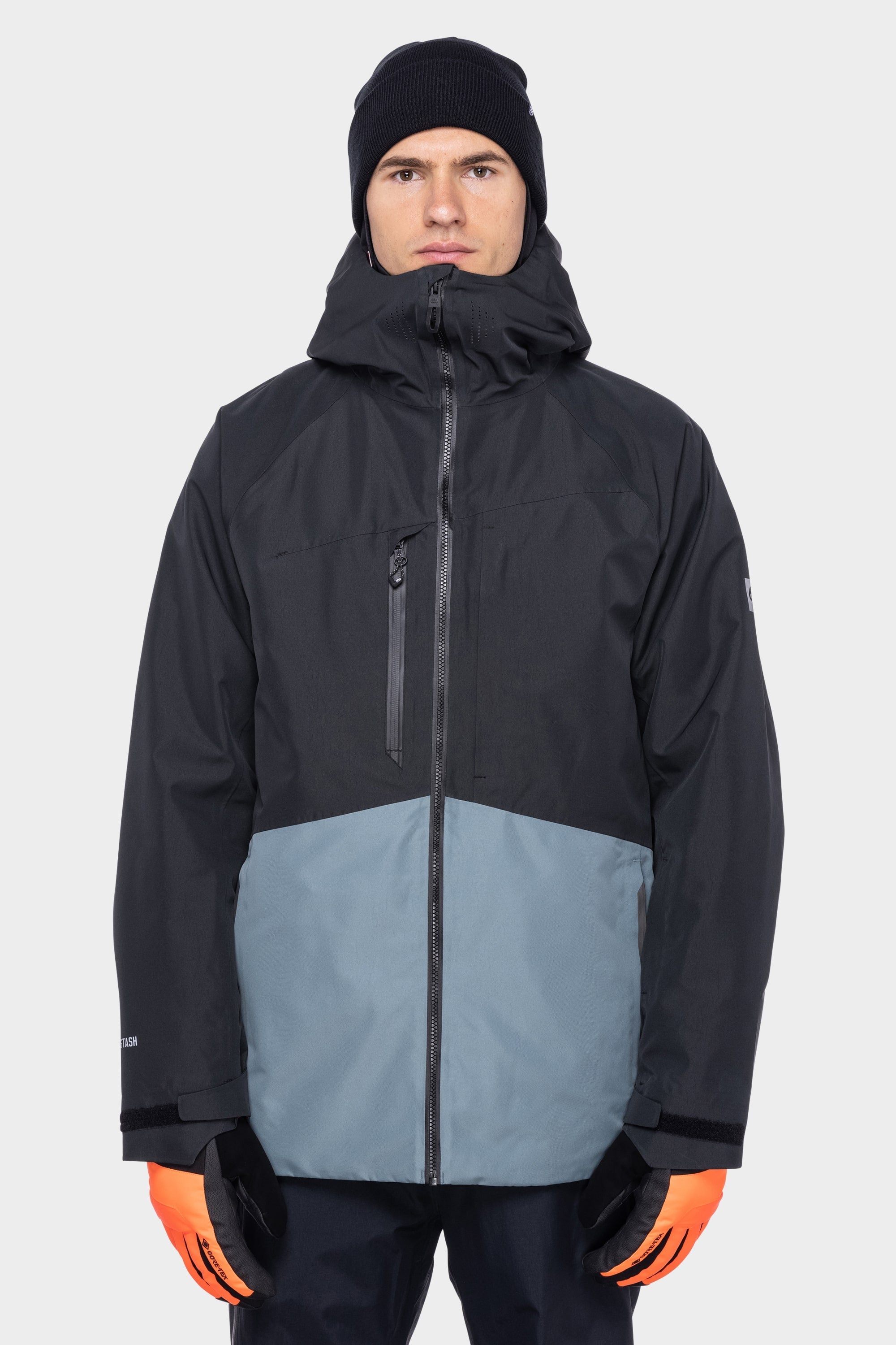 A person wears a 686 Men's Hydrastash Reserve Insulated Jacket—black and gray, hooded, waterproof polyester—paired with a black beanie and bright orange inner gloves, standing facing forward against a plain light gray background.