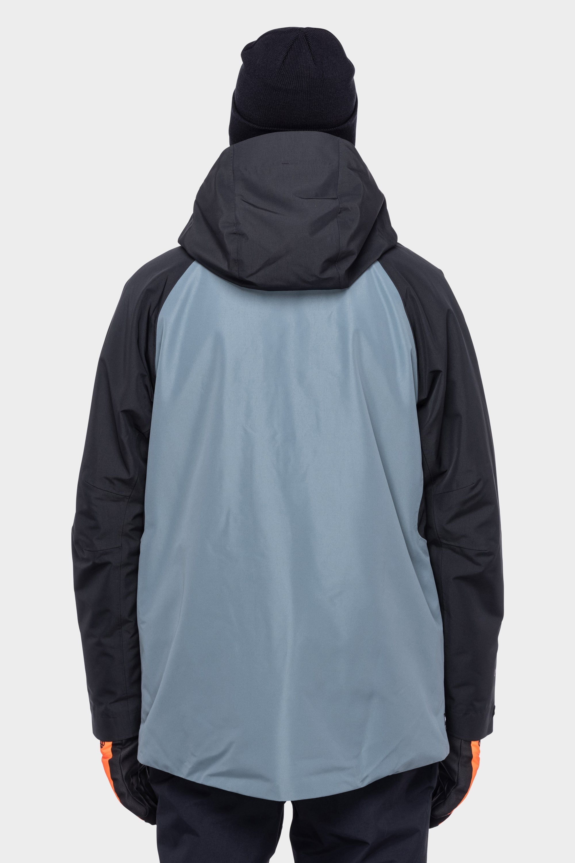 A person wearing a black beanie, orange gloves, black pants, and the 686 Men's Hydrastash Reserve Insulated Jacket in blue and black stands with their back to the camera against a plain light background.