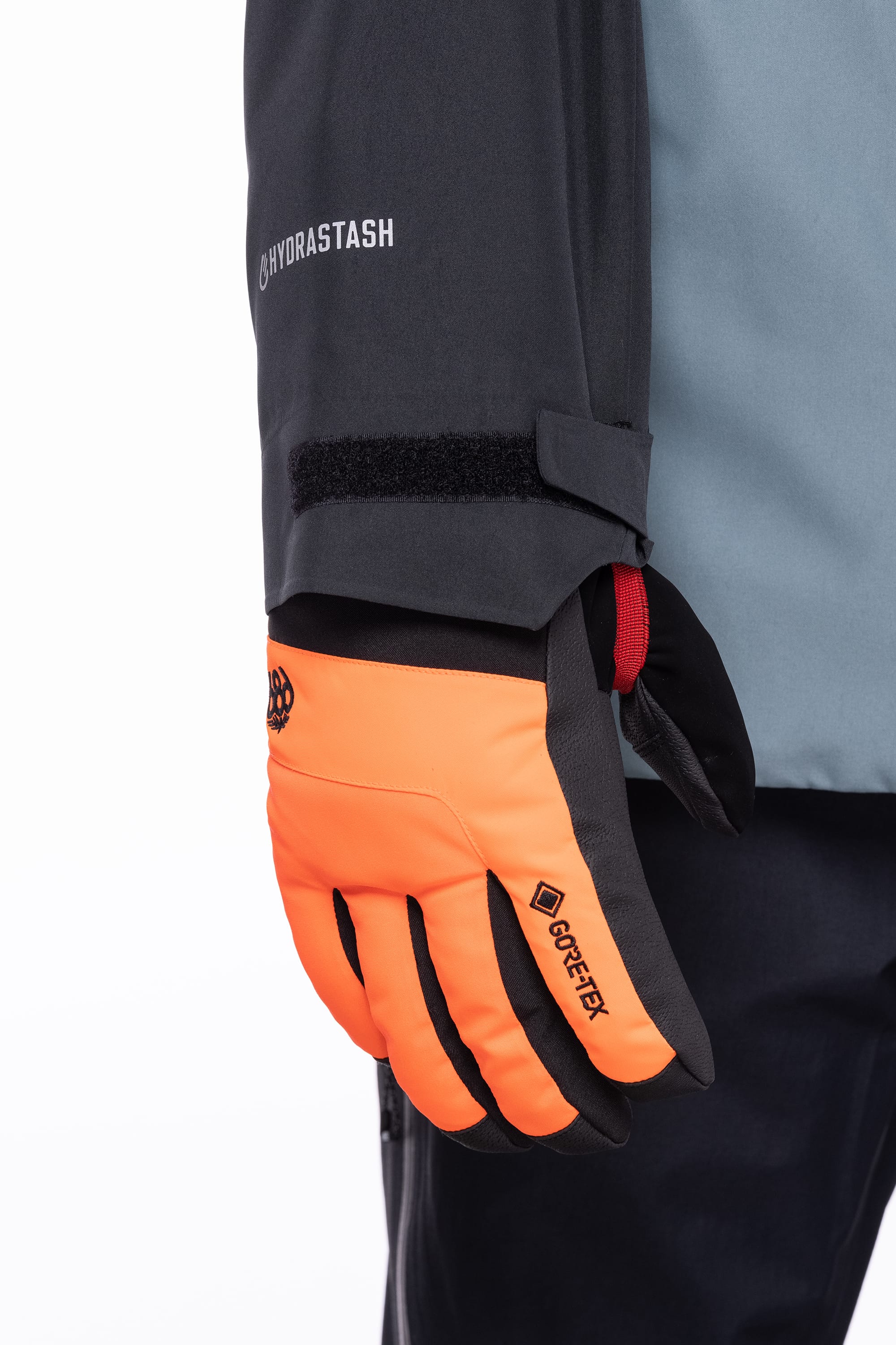 A person wearing a 686 Men's Hydrastash Reserve Insulated Jacket in gray and black with HYDRASTASH branding on the sleeve, paired with bright orange GORE-TEX gloves and black pants.