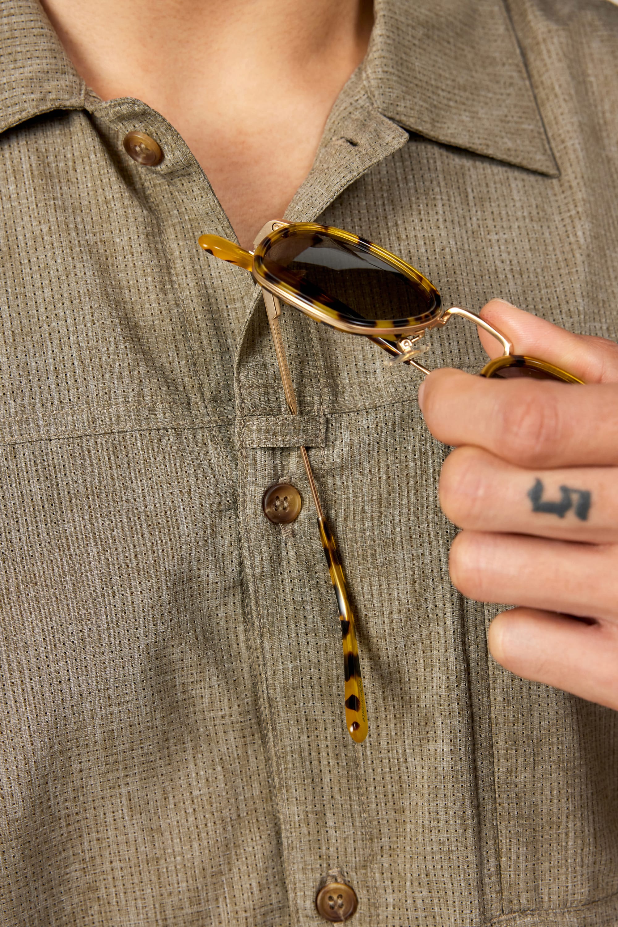 A person in a beige 686 Mens Canopy Perforated Button Up shirt holds tortoiseshell sunglasses, displaying a 13 tattoo on their ring finger.