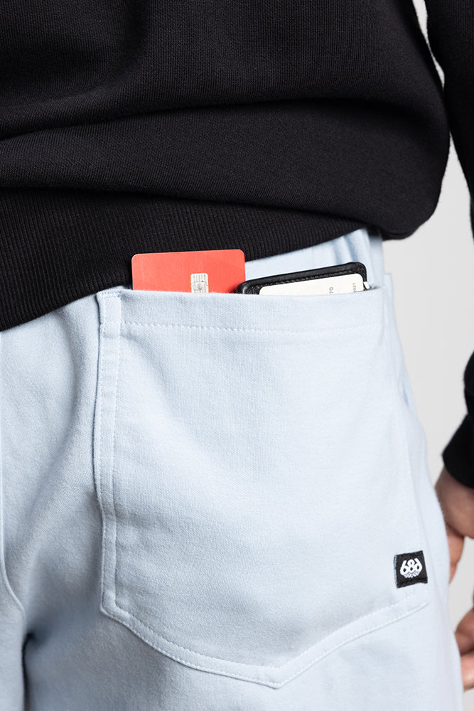 A close-up of a person wearing 686 Mens Everywhere Double Knit Shorts in light blue, paired with a black sweater. A red card and smartphone are partially visible in the front pocket.