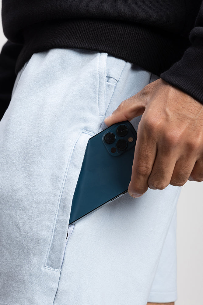 A person wearing 686 Mens Everywhere Double Knit Short in light blue and a black top is putting a dark blue smartphone into the shorts’ side pocket.